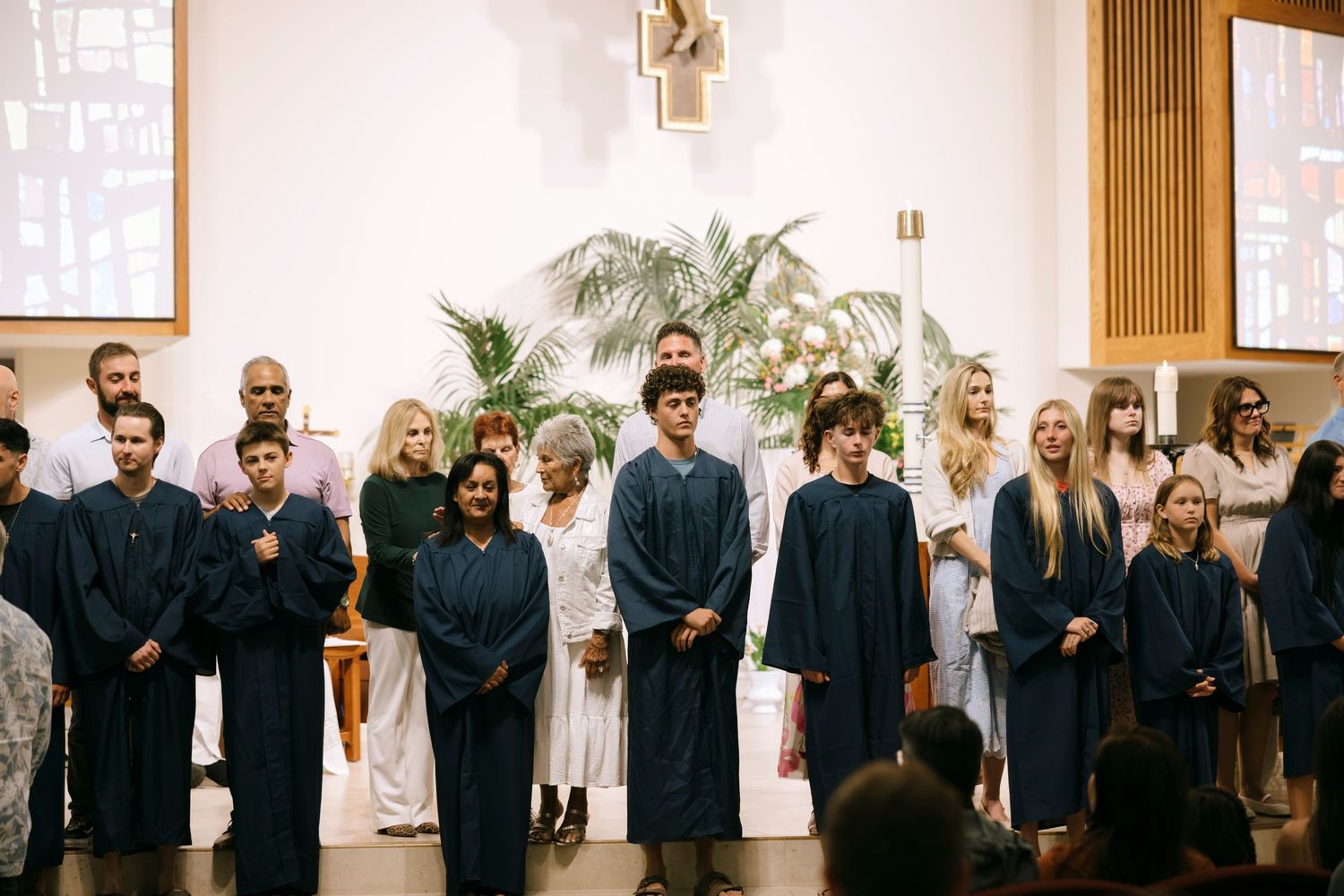 Students in blue graduation gowns stand at a church ceremony with white-robed attendees and floral arrangements.