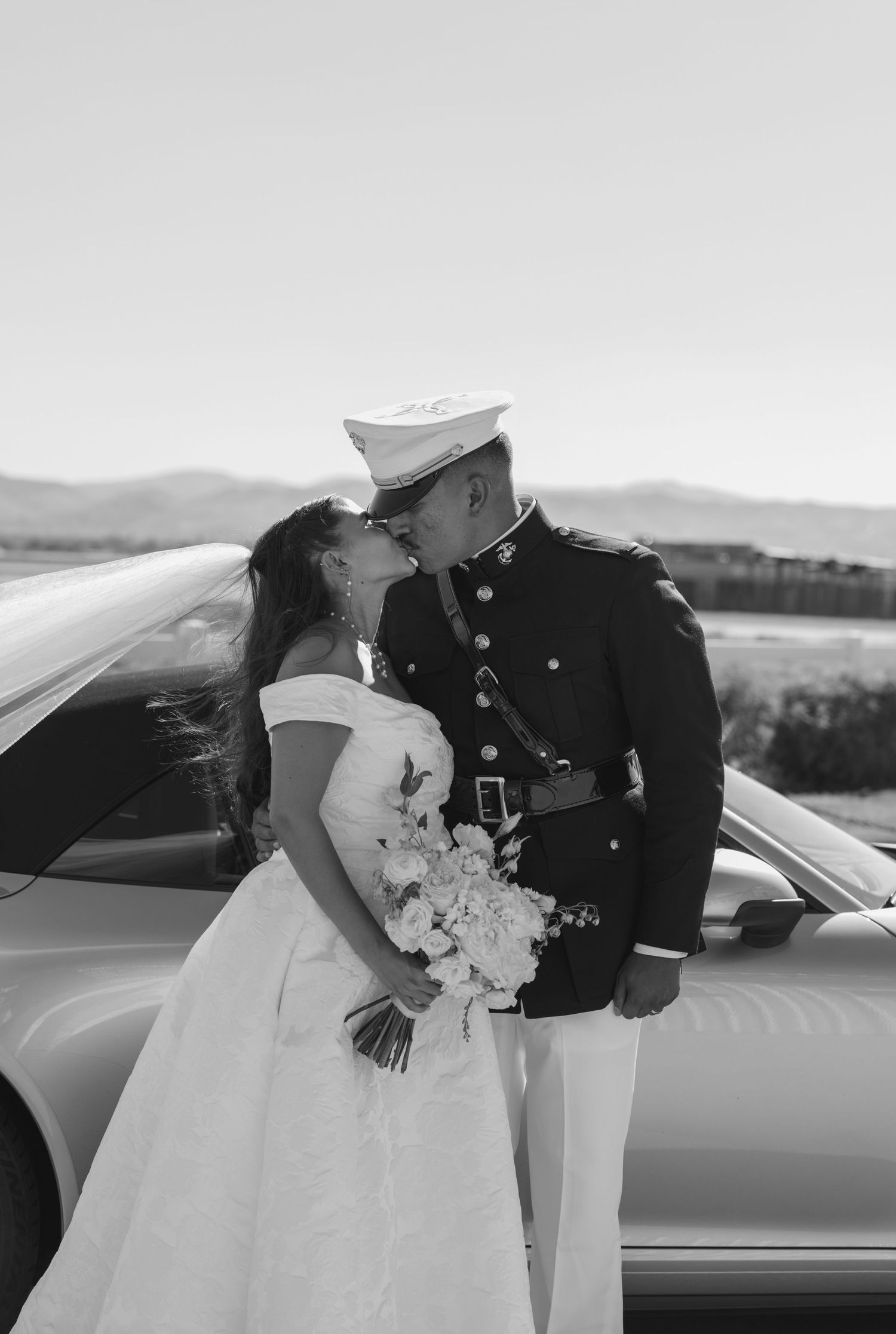 Black and white portrait of a military couple with their wedding attire by a car.