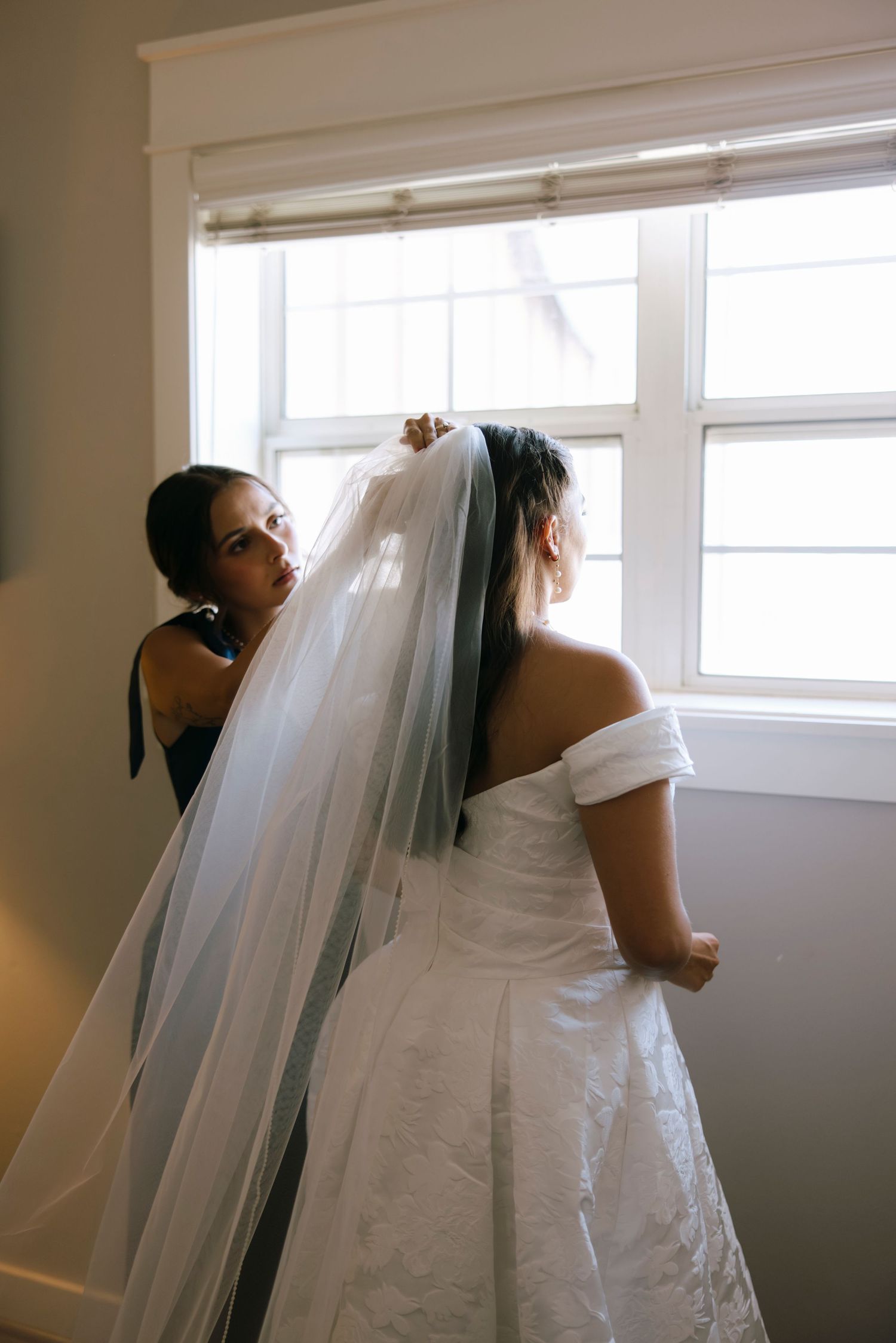 A bride adjusts her veil near bright windows in a minimalist white room.