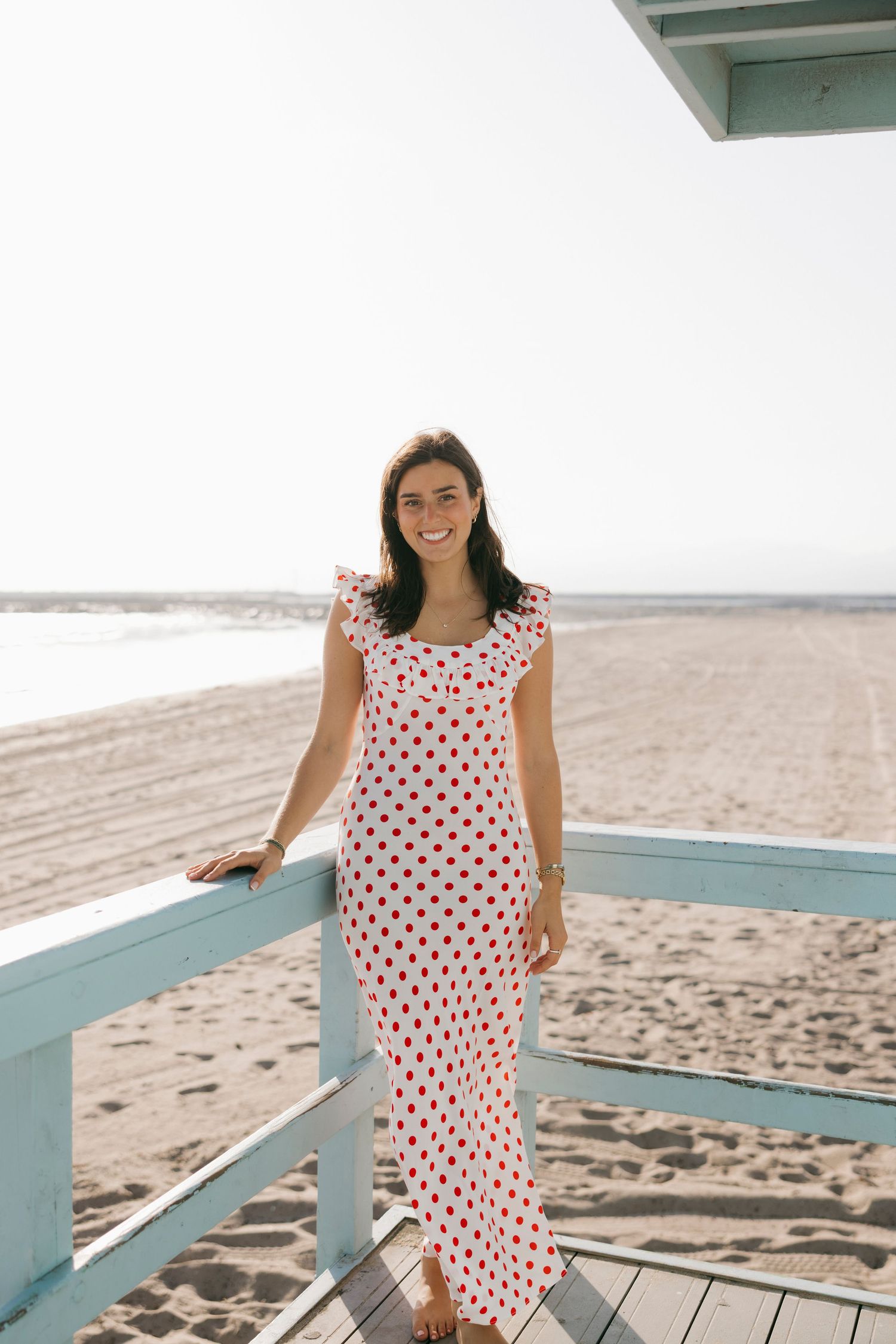 Young woman in polka dot maxi dress smiling on a beach lifeguard stand.
