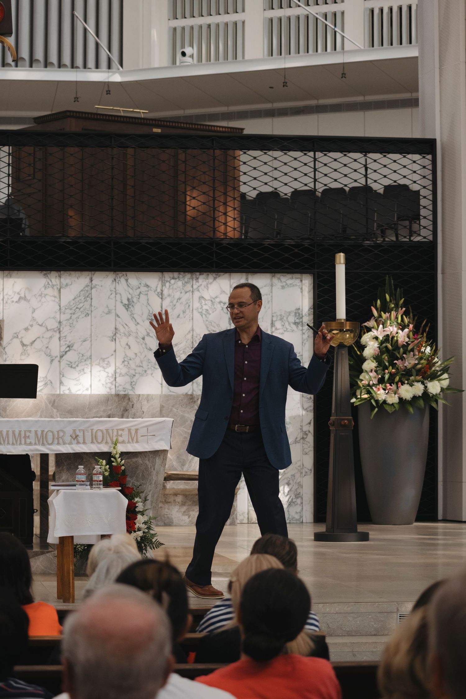 A speaker in a blue blazer gestures while presenting on stage at a church or conference venue with floral decorations.