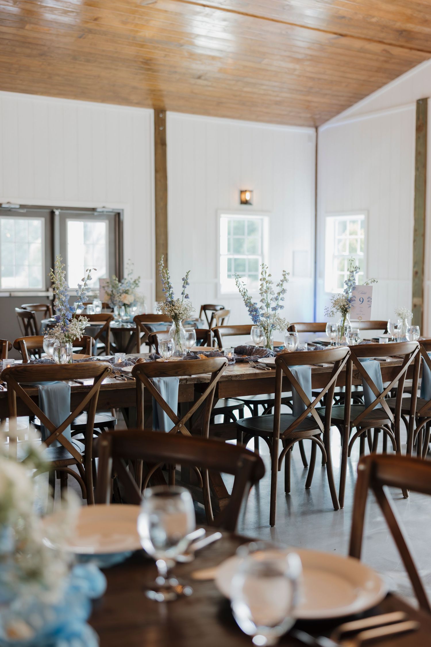 Rustic barn wedding reception with wooden cross-back chairs, long tables, blue floral centerpieces, and elegant place settings.