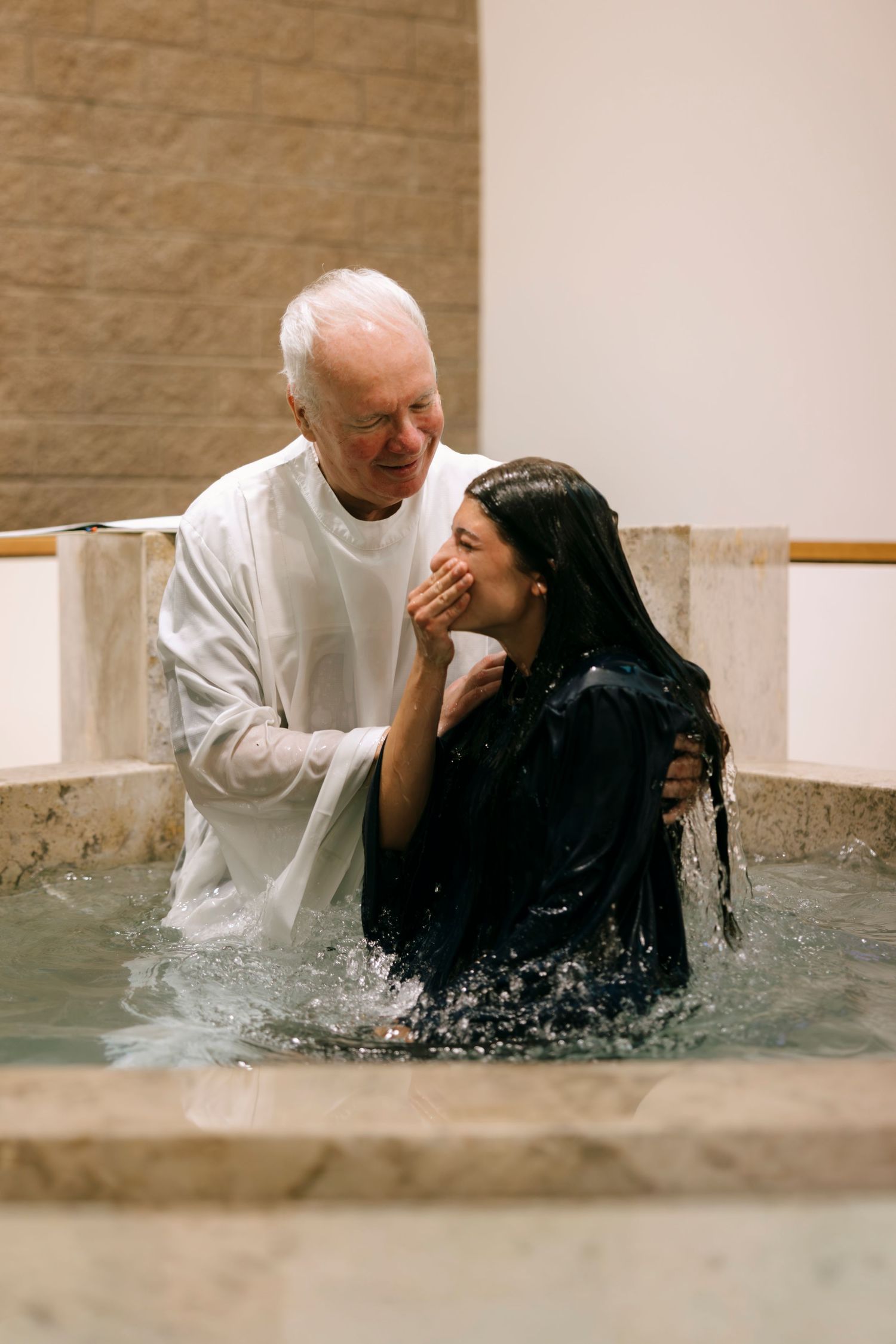 Elderly pastor baptizing a young woman in a church baptismal pool as water drips from her hair.