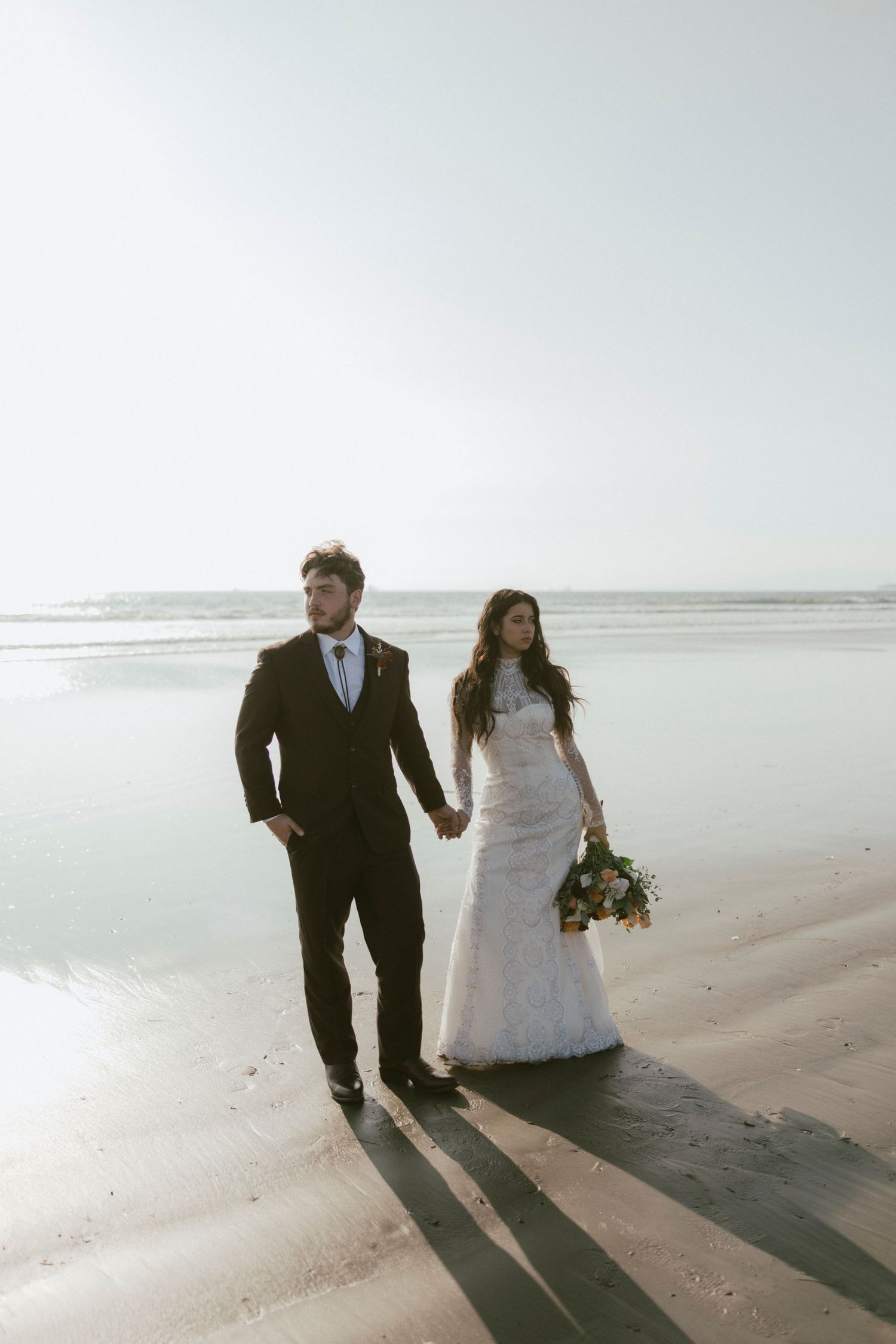 Bride and groom walking along shoreline casting long shadows on wet sand.