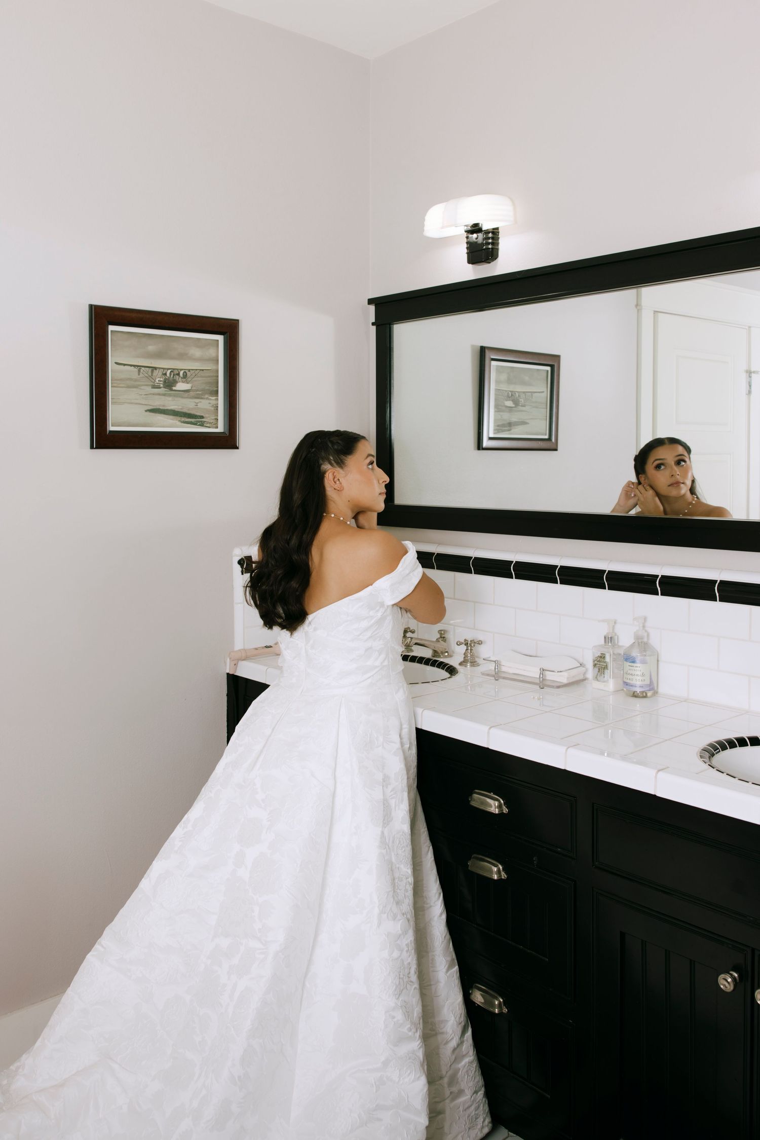 A bride in a white wedding dress stands near a dark bathroom vanity with framed artwork on the wall.