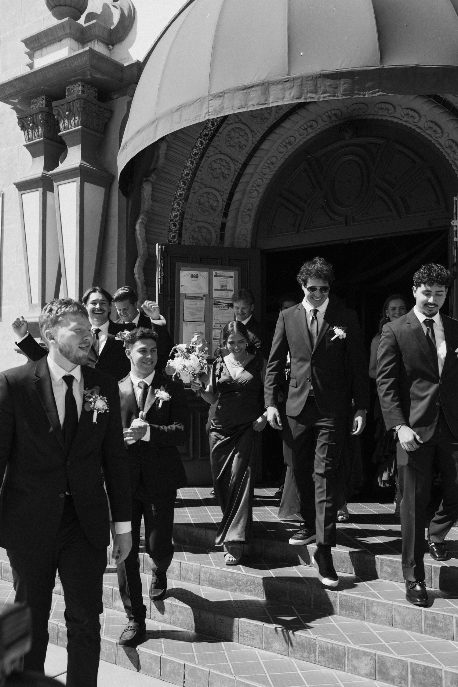 Groomsmen in dark suits pose on brick steps outside a church entrance.