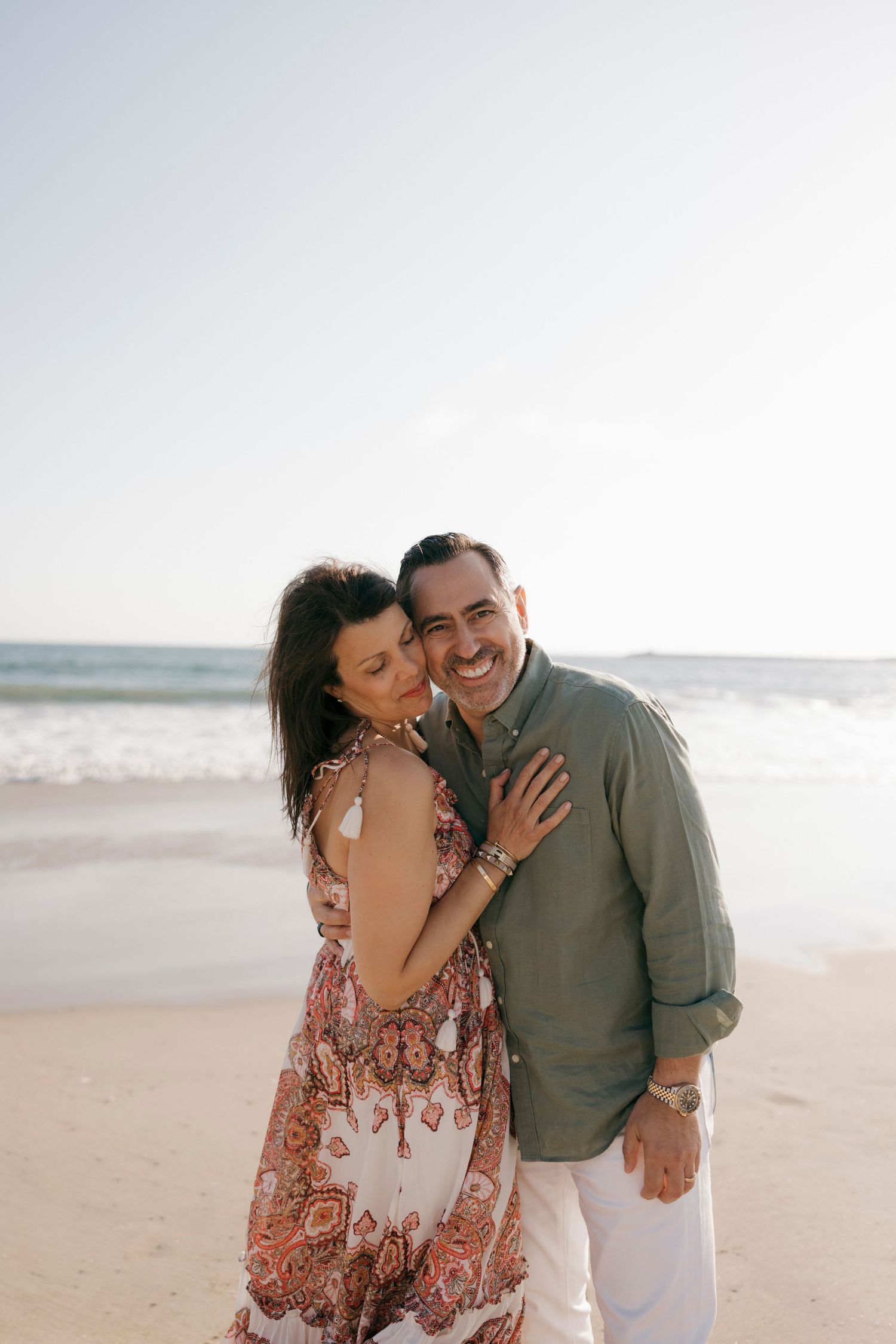 Couple embracing and smiling on a sunny beach with ocean in background.