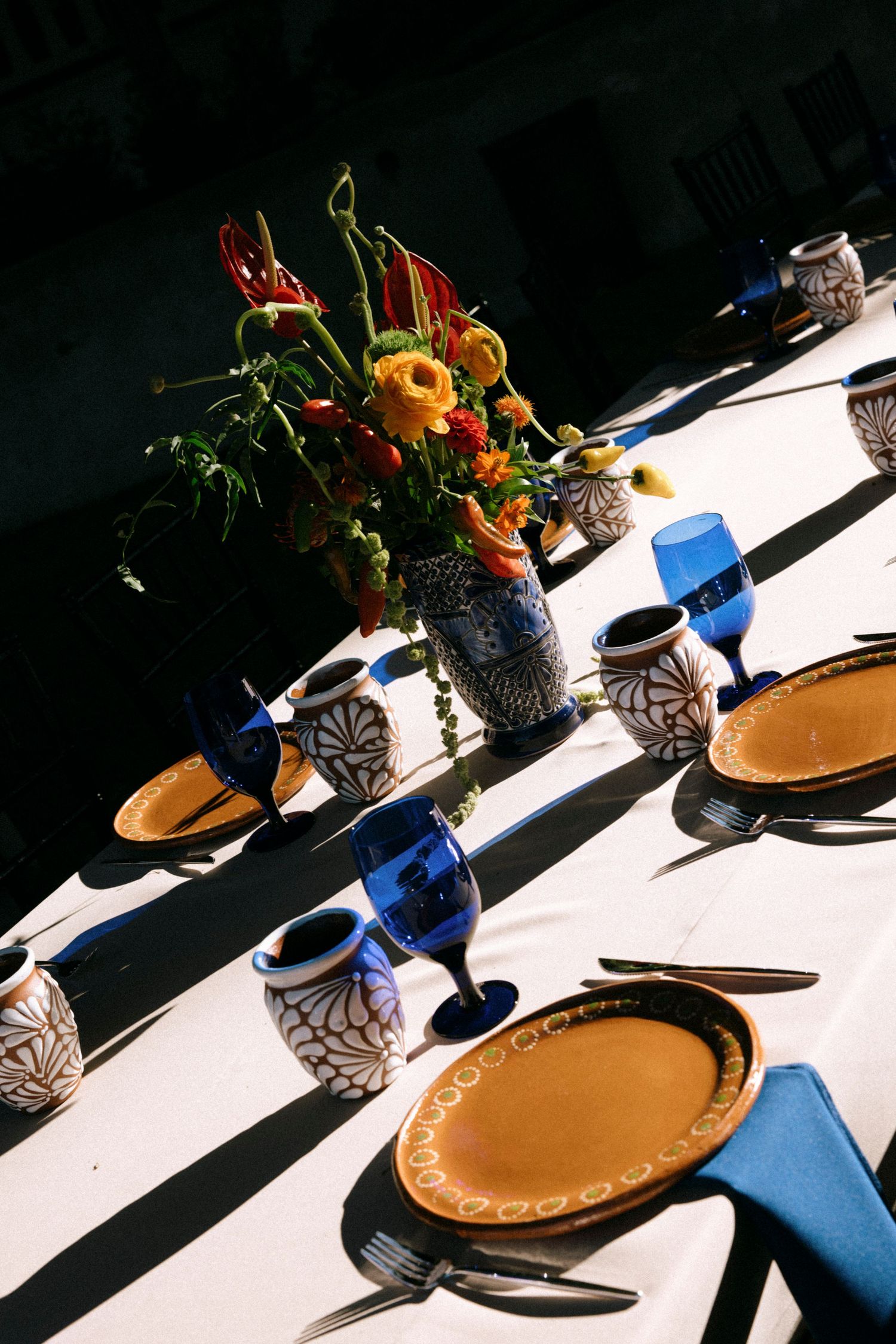 Elegant table setting with orange flowers in vases and blue glassware creating dramatic shadows.