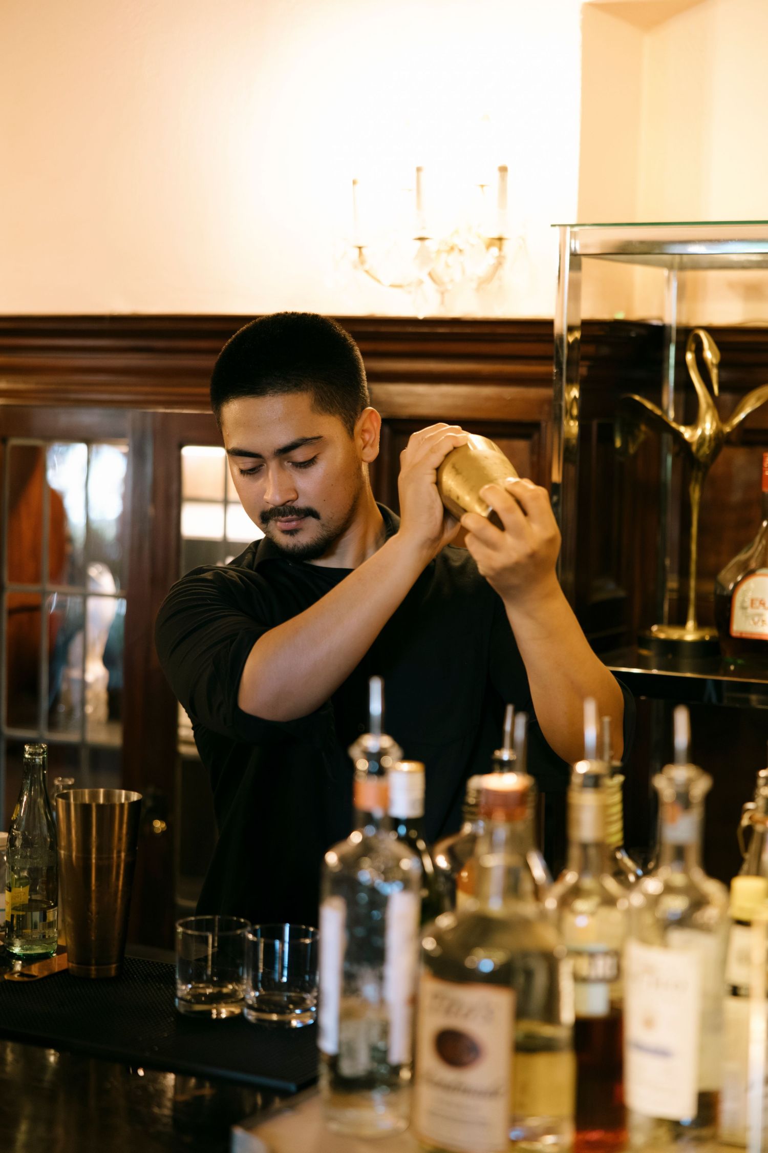 Bartender preparing drinks behind a dark wooden bar with bottles and glassware.