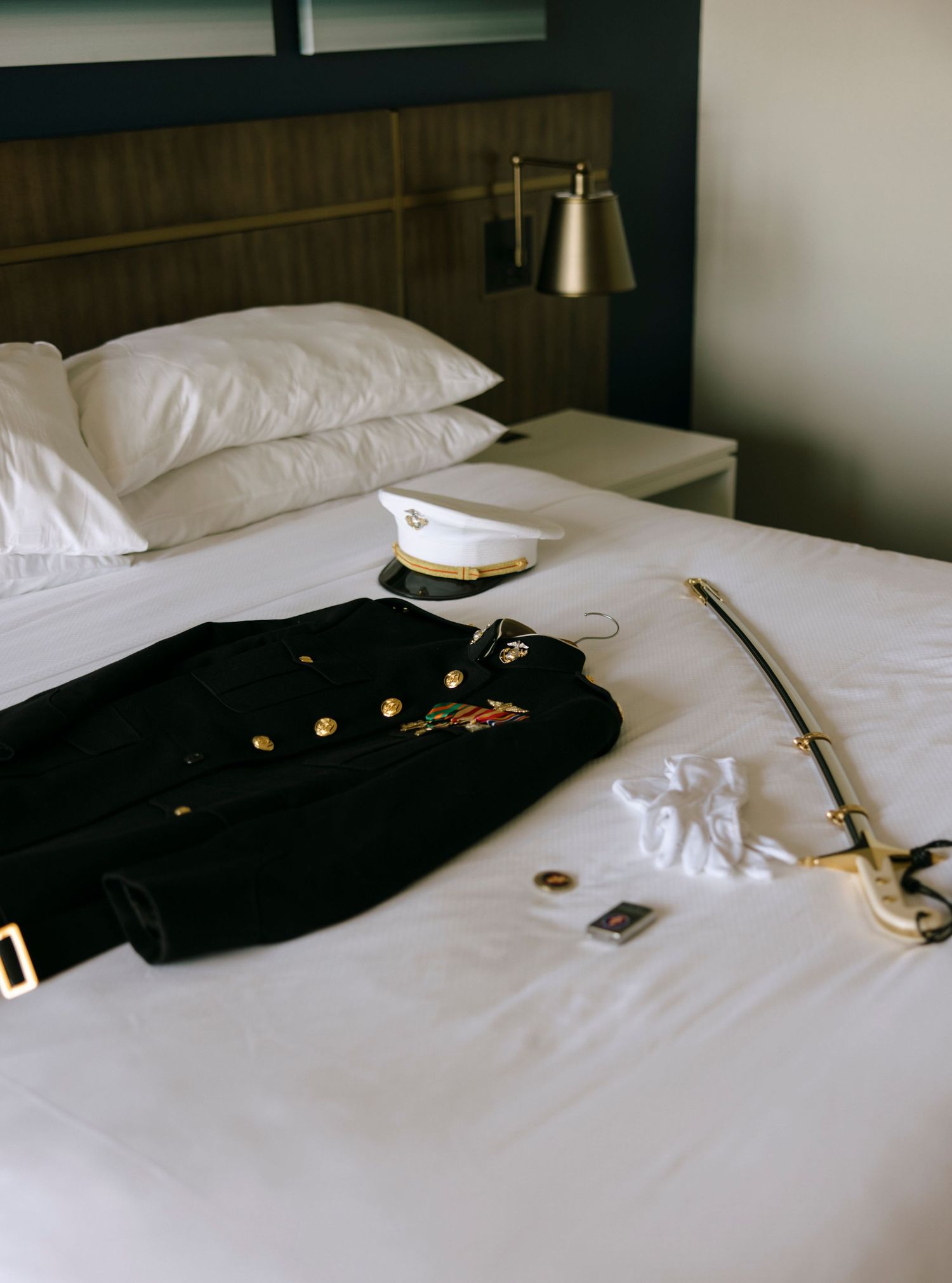 Military dress uniform jacket, white officer cap, sword, gloves, and medals laid out on a white hotel bed.
