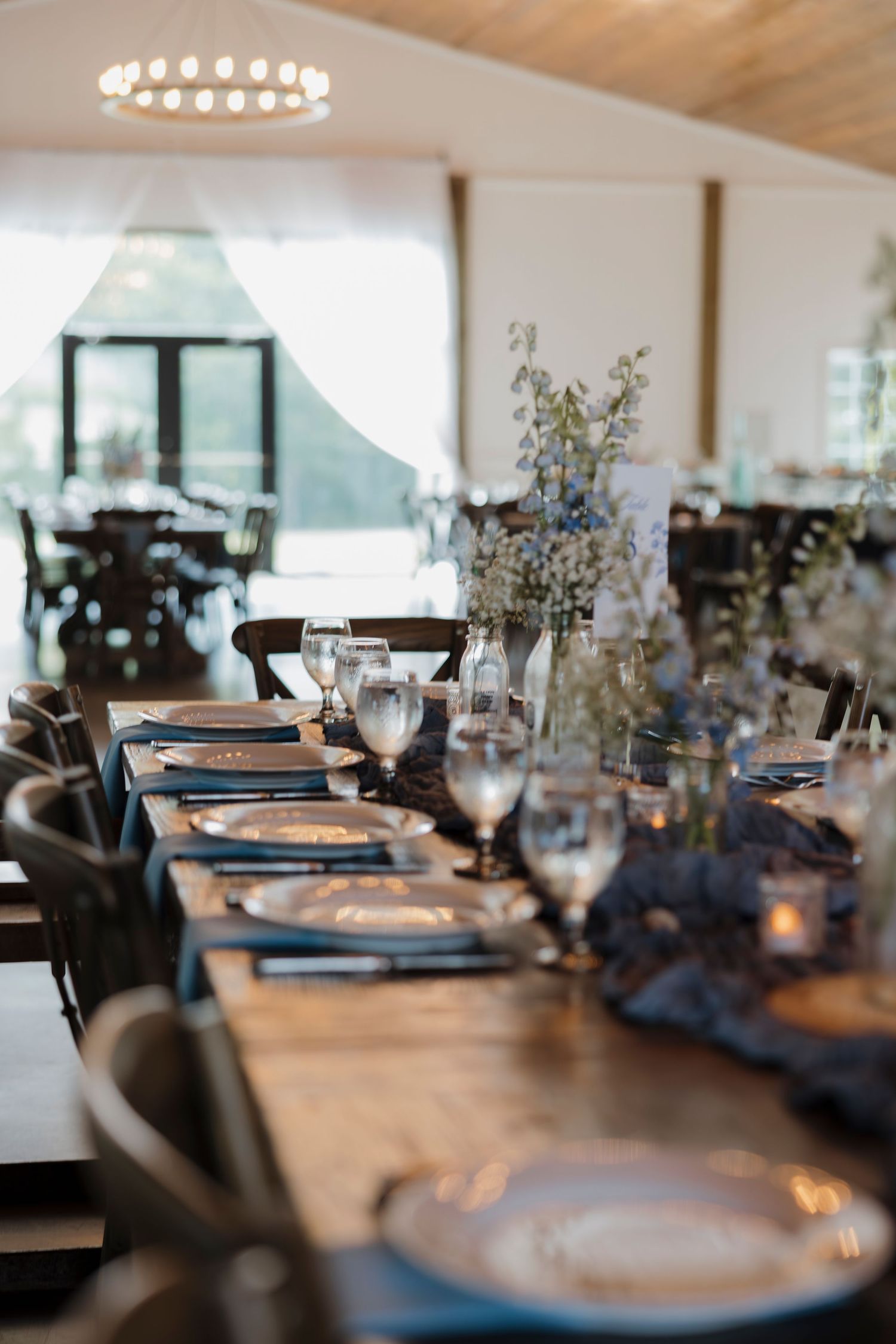 Elegant rustic wedding reception table set with navy napkins, crystal glasses, silver chargers, and floral centerpieces.