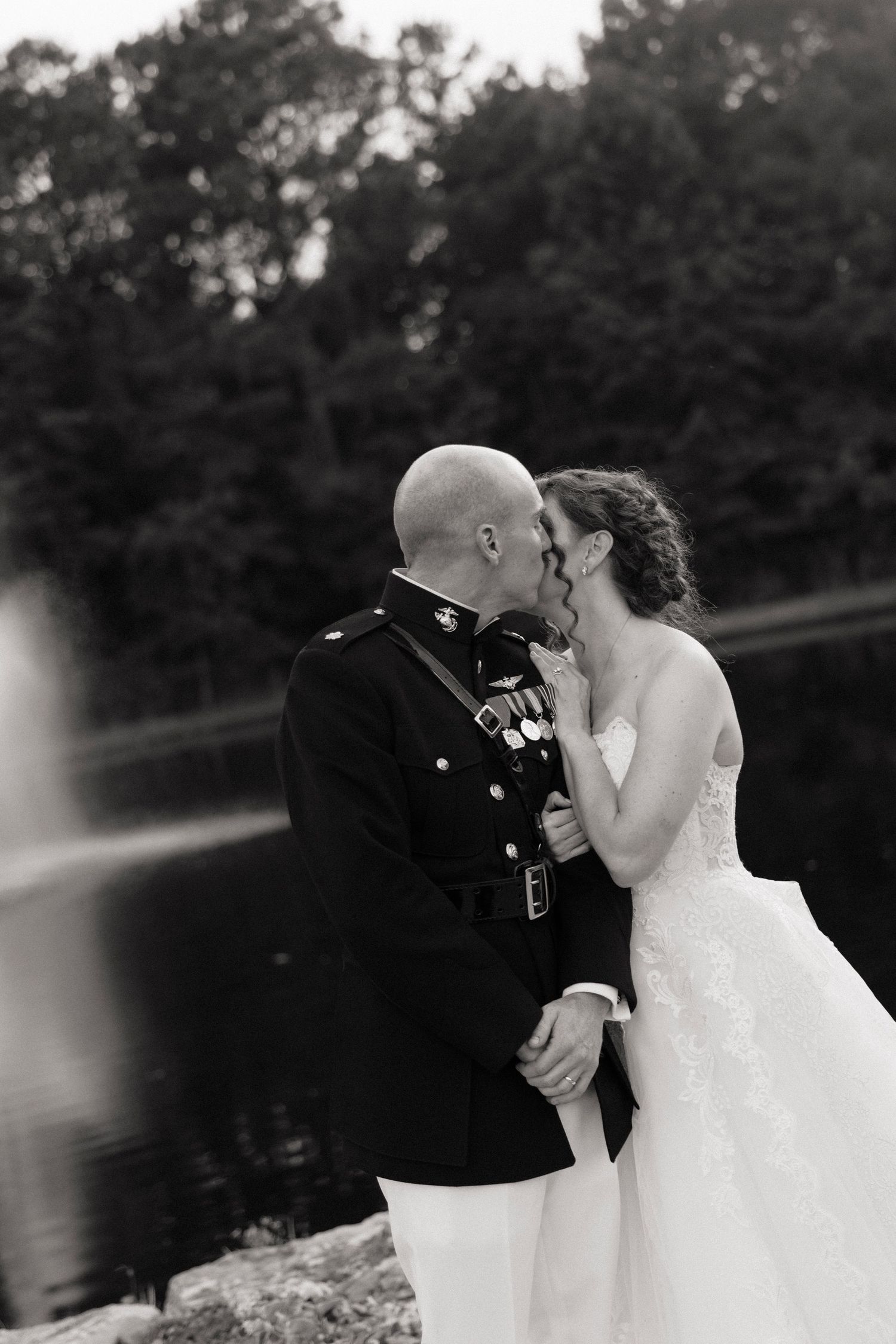 Black and white photo of a military groom in uniform kissing his bride in a strapless wedding gown near a serene lake.