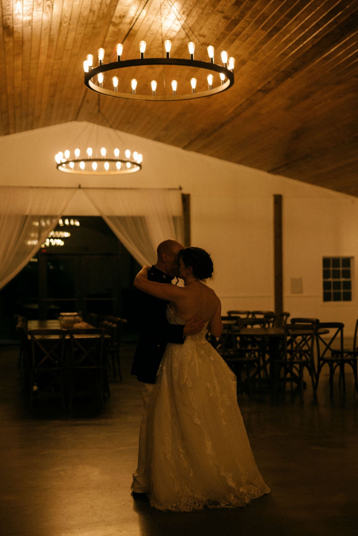 Bride and groom share first dance under a circular chandelier in a warmly lit rustic wedding venue.