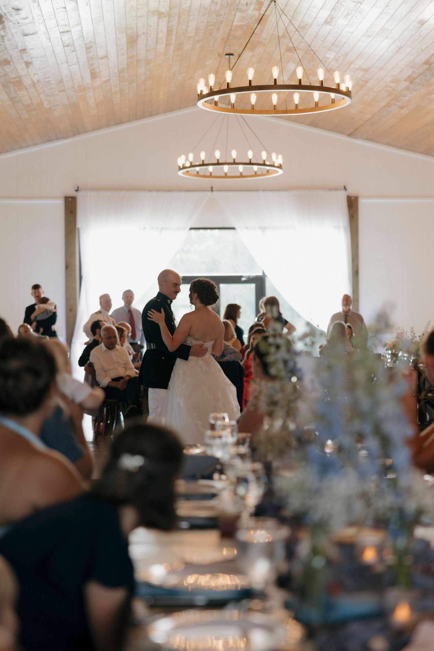 Bride and groom share first dance in elegant barn venue with candle chandelier and guests seated at decorated tables.