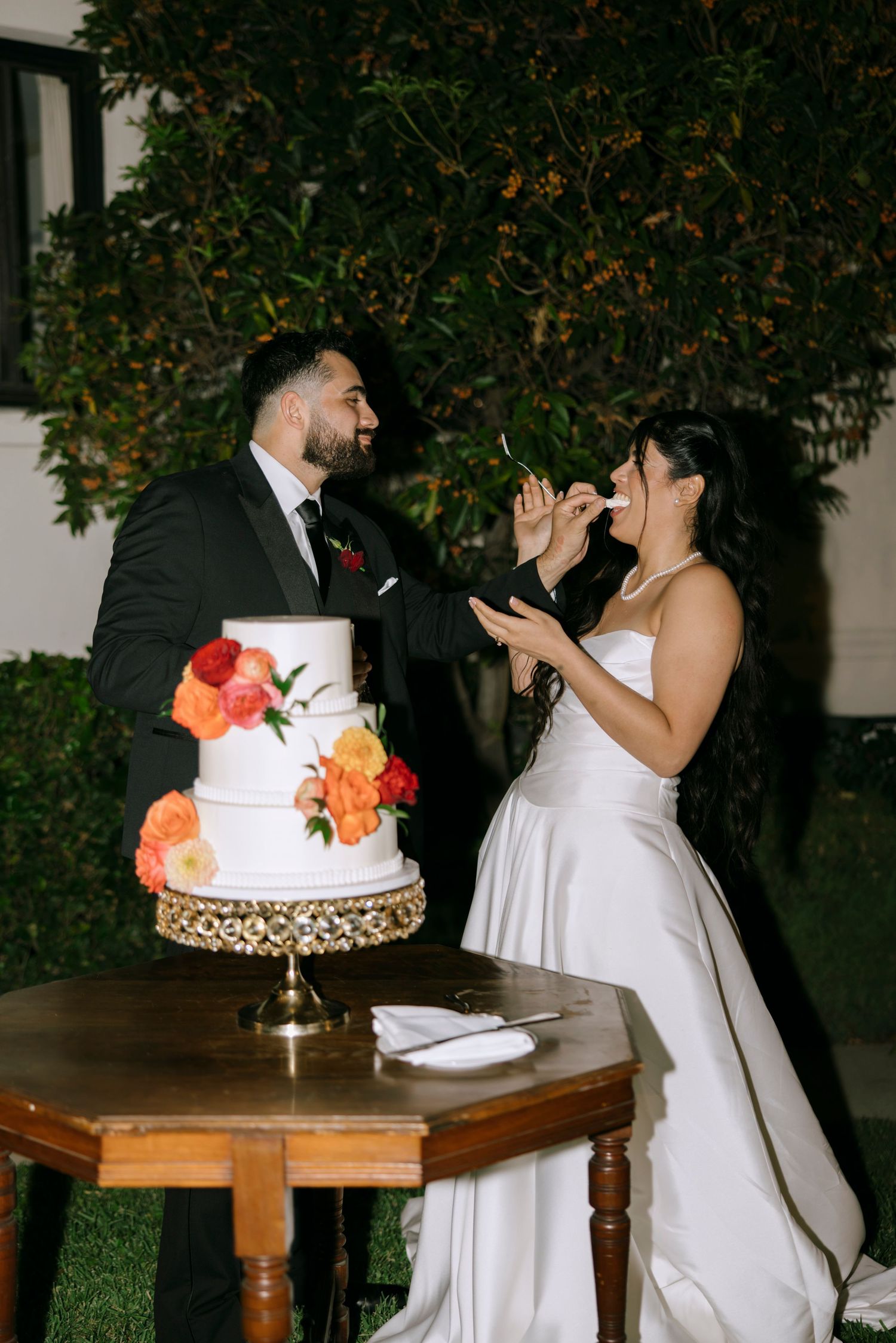 A couple laughs together while standing next to their elegant three-tiered wedding cake.