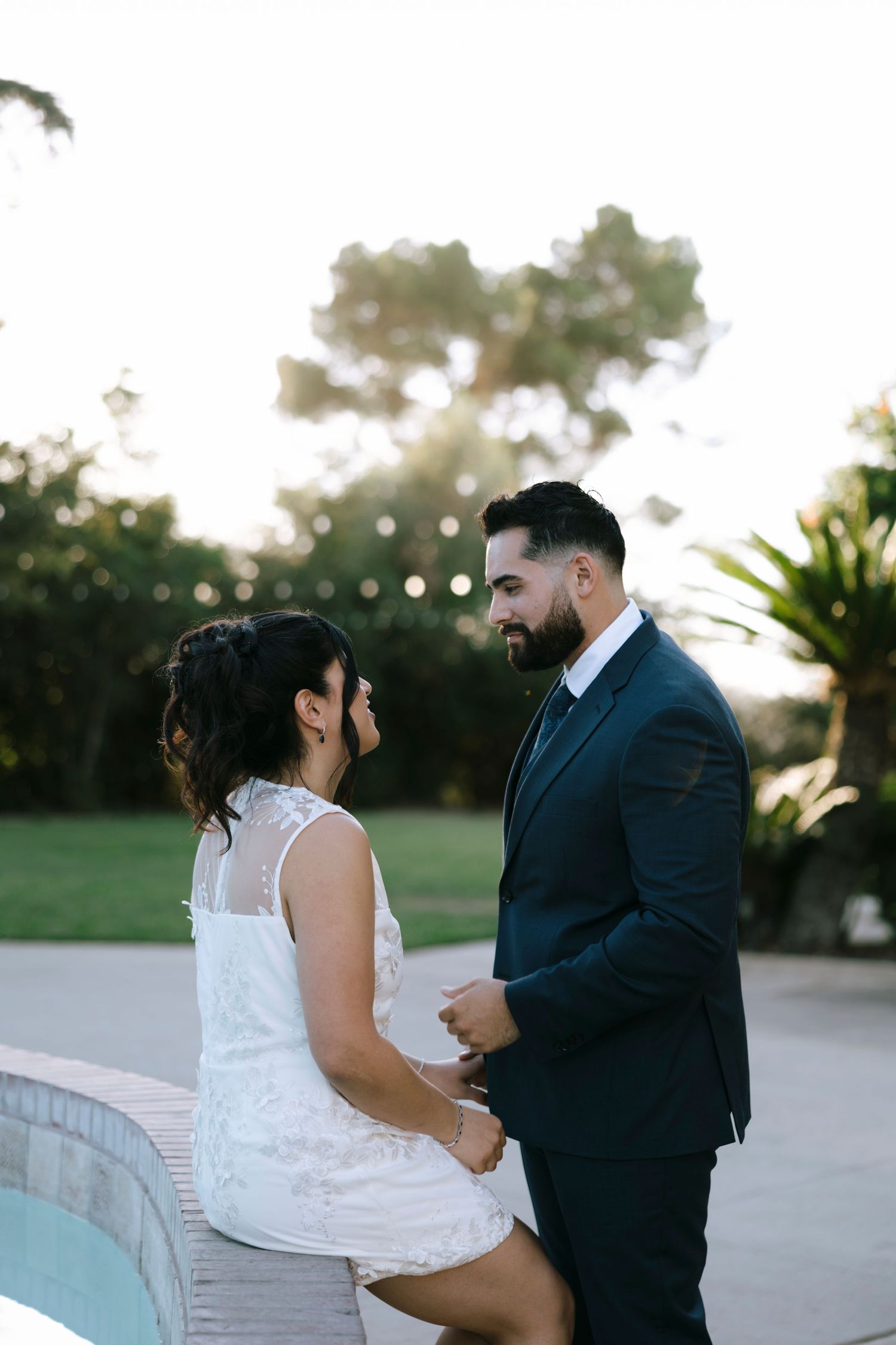 A romantic couple shares an intimate moment by a pool wall at sunset.