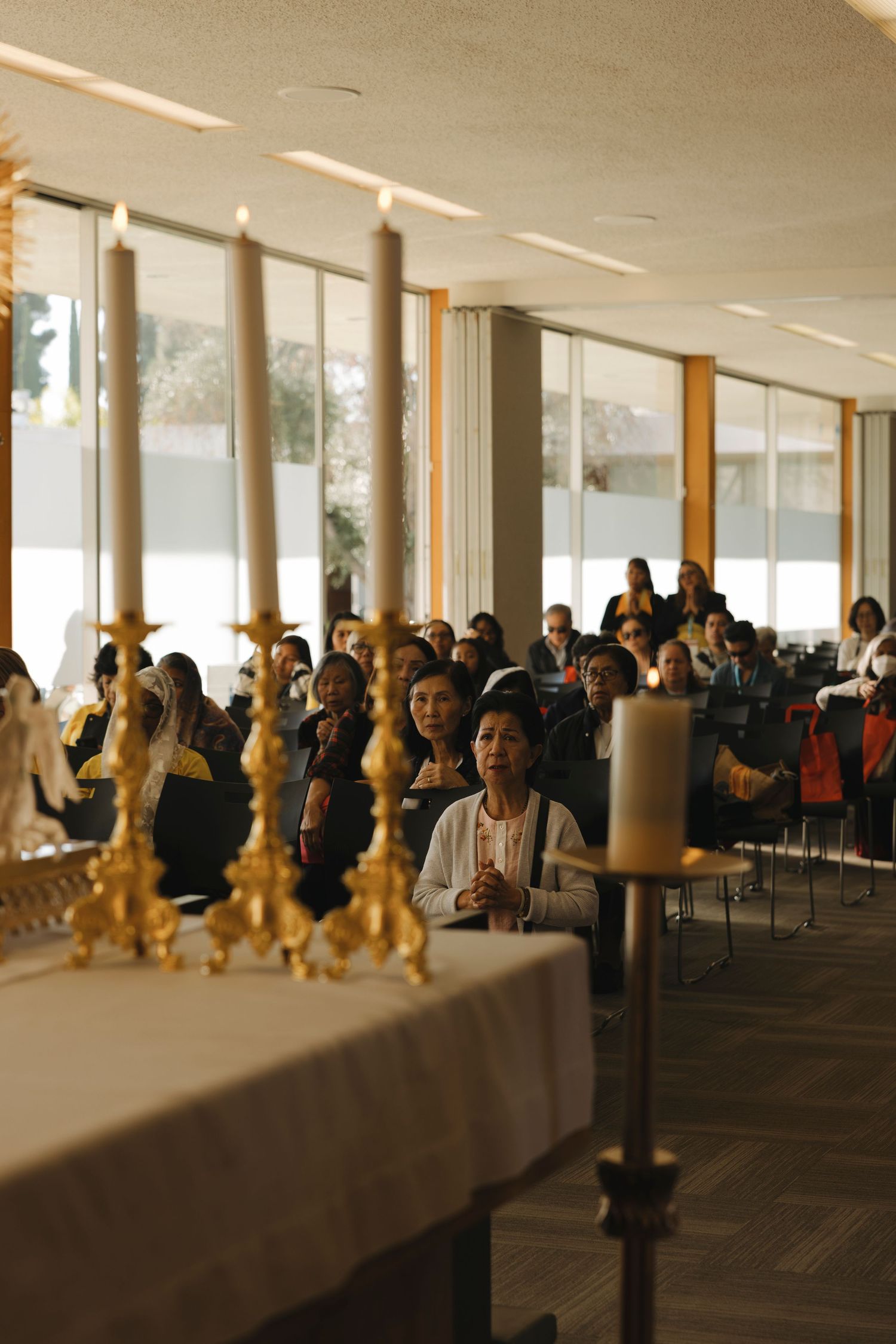 Crowd seated in a bright room with floor-to-ceiling windows and wooden columns.