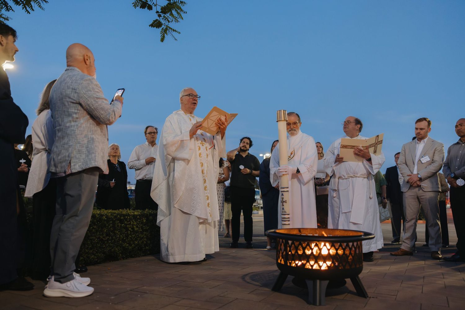 Catholic priests in white vestments perform an outdoor evening blessing ceremony around a fire pit at dusk.