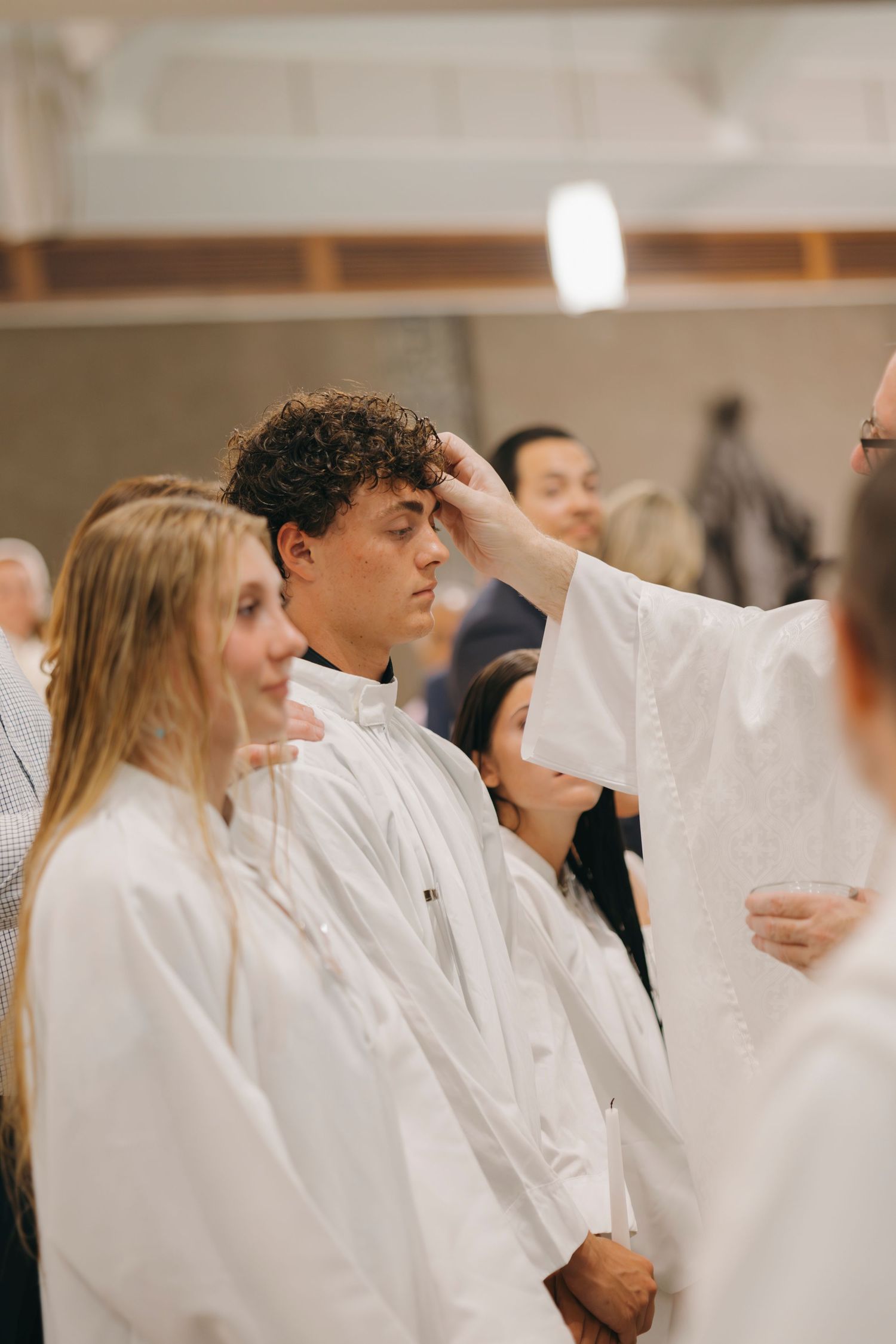 Catholic priest anointing young man's forehead with chrism oil during Confirmation sacrament ceremony.
