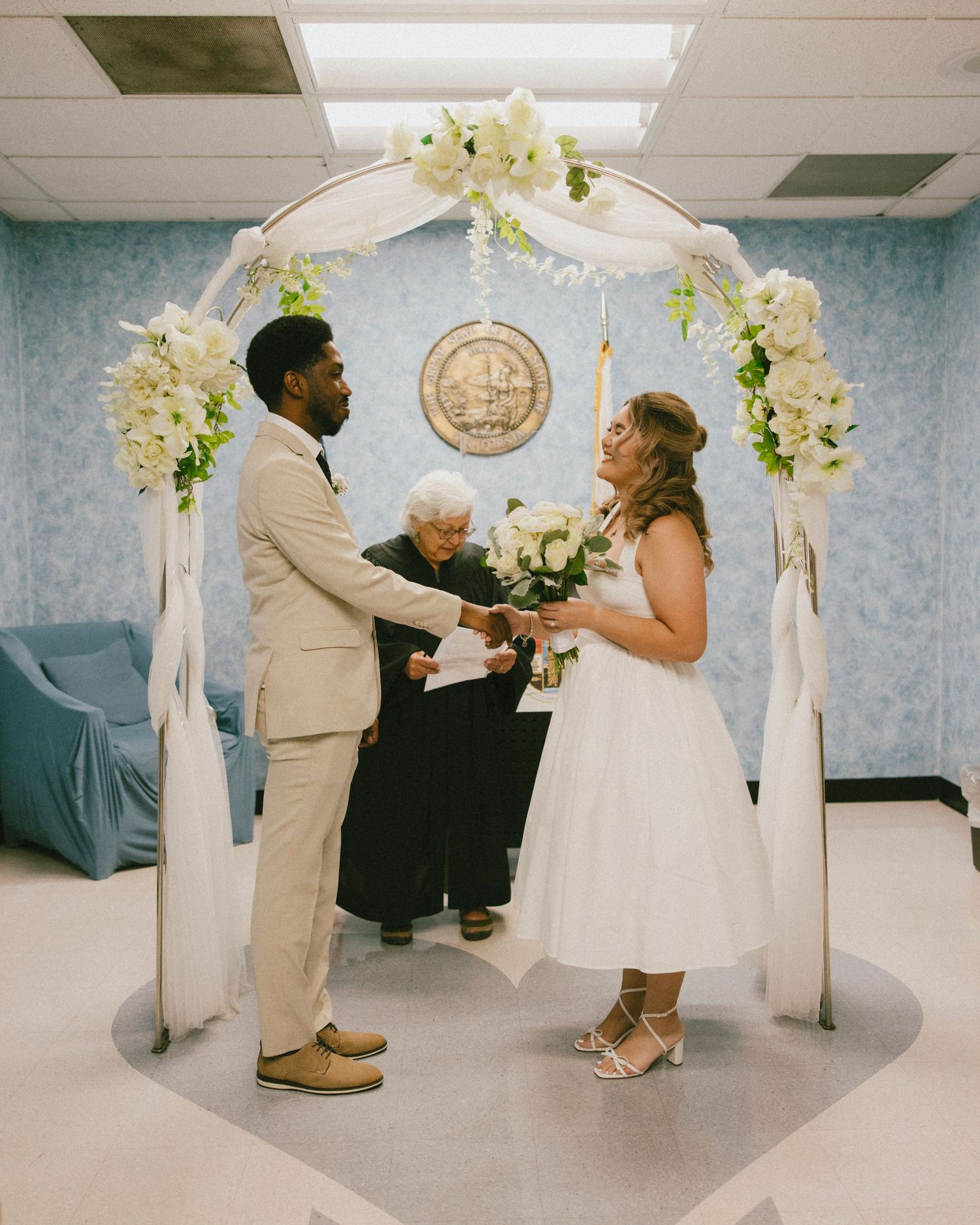 A wedding ceremony under a white floral arch with the couple exchanging vows.