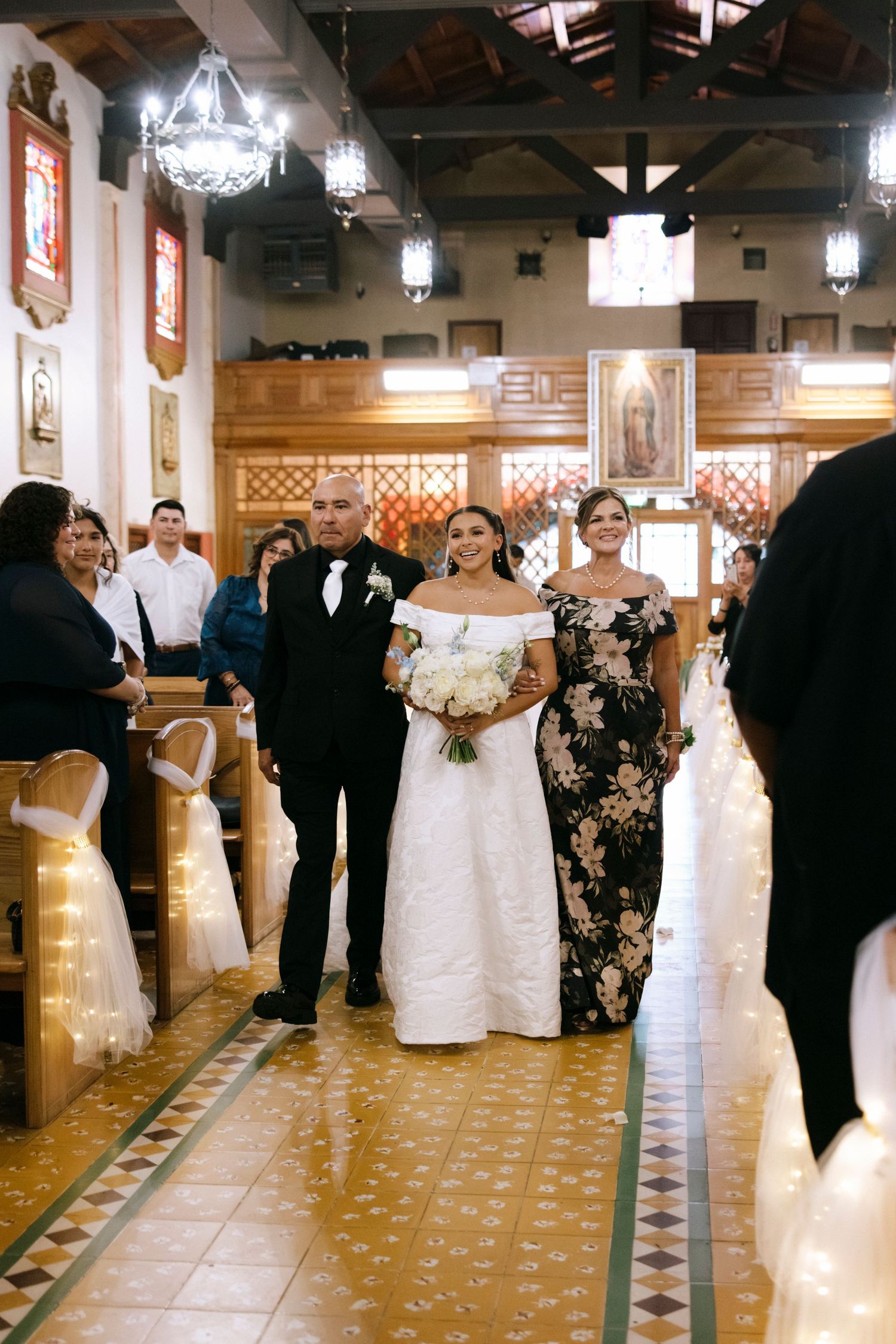 Wedding party members walk down the aisle of an ornately decorated church with tiled floors.