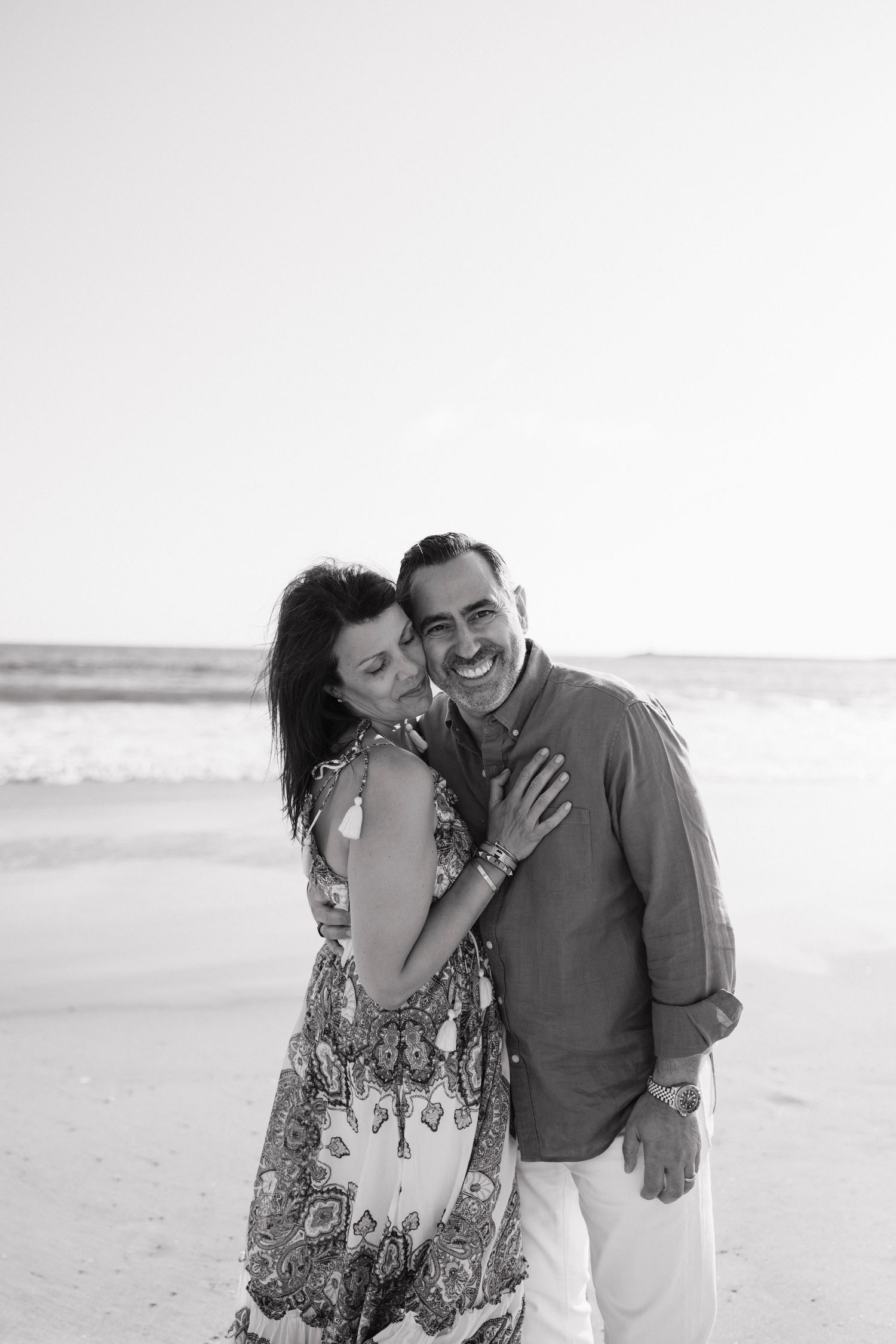 Black and white photo of a happy couple embracing on a beach, woman in patterned dress leaning on smiling man.