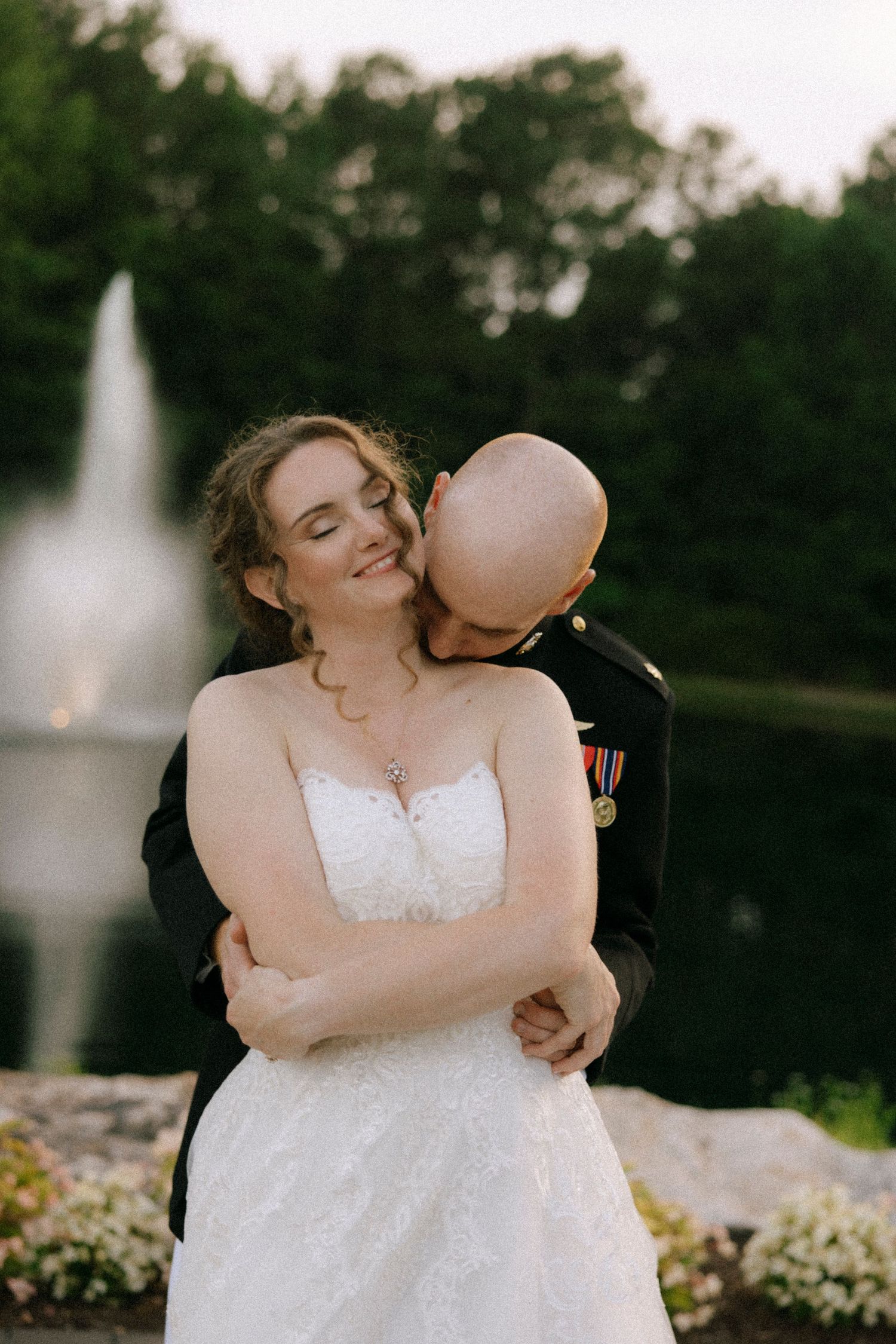 A Marine groom in dress uniform embraces and kisses his smiling bride in a white gown near a garden fountain.