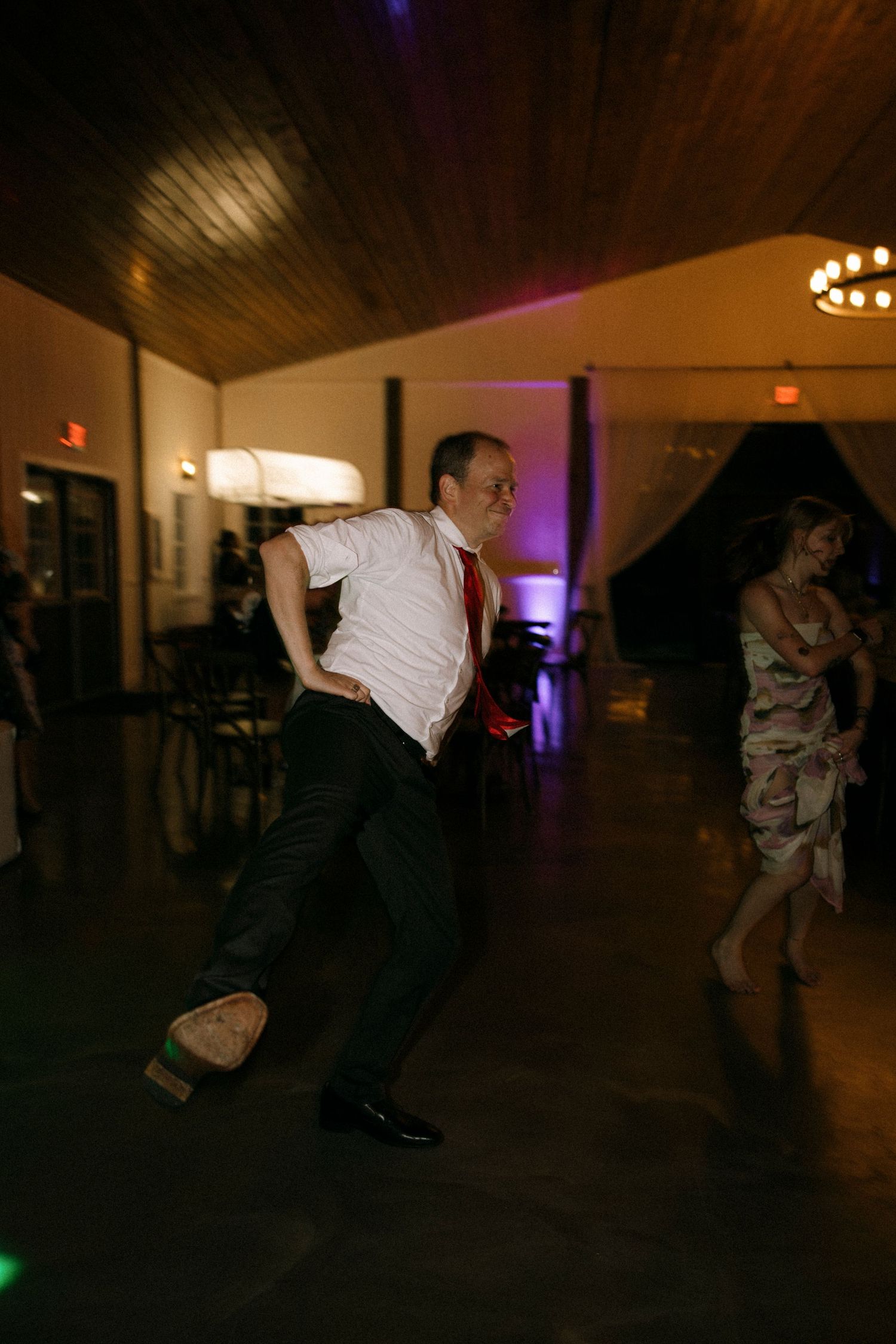Man in white shirt and red tie dancing energetically at an indoor wedding reception venue with purple ambient lighting.