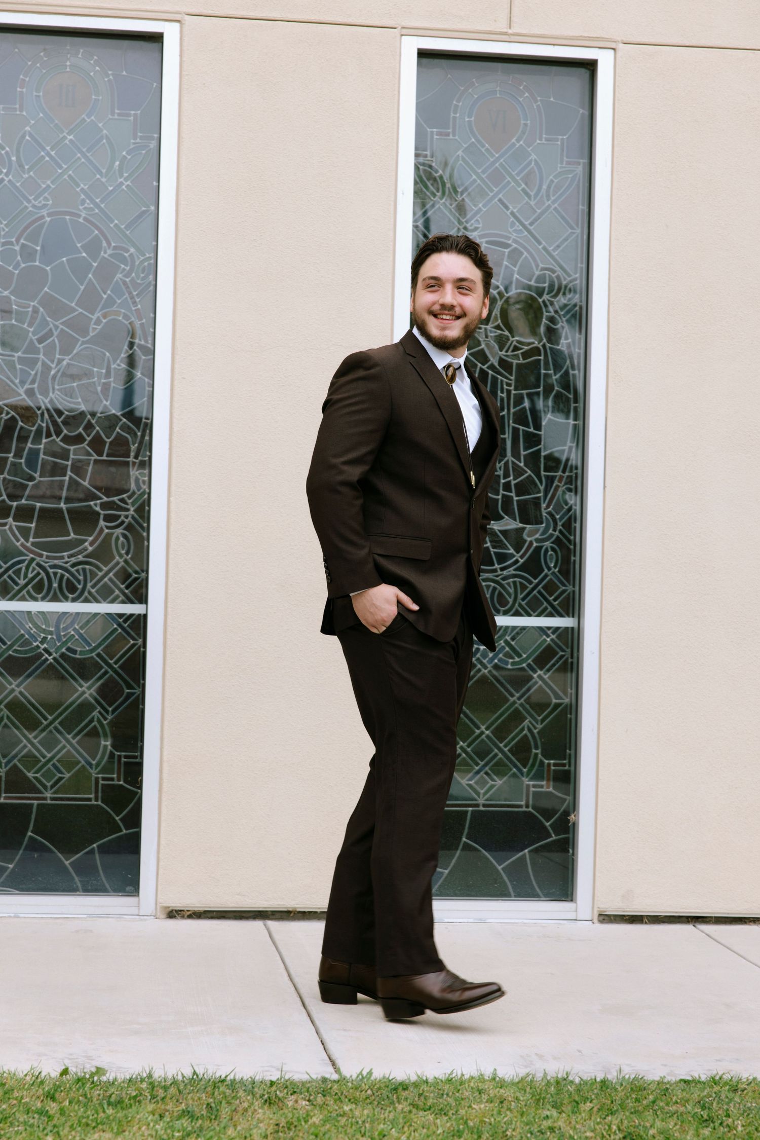 Professional in brown suit poses casually against wall with decorative glass panels.