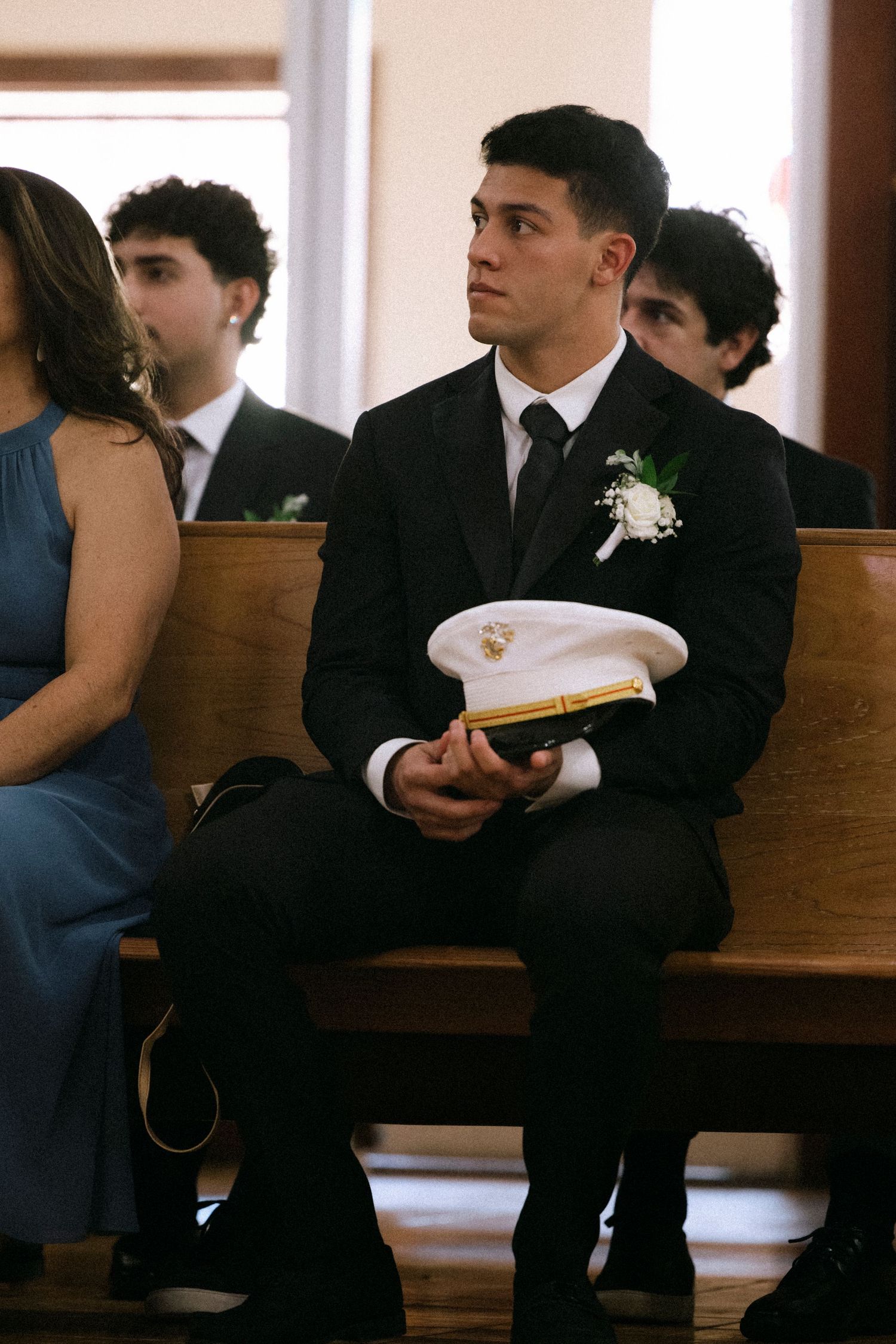 A formal attendee in a black suit sits in a church pew holding a military service cap.