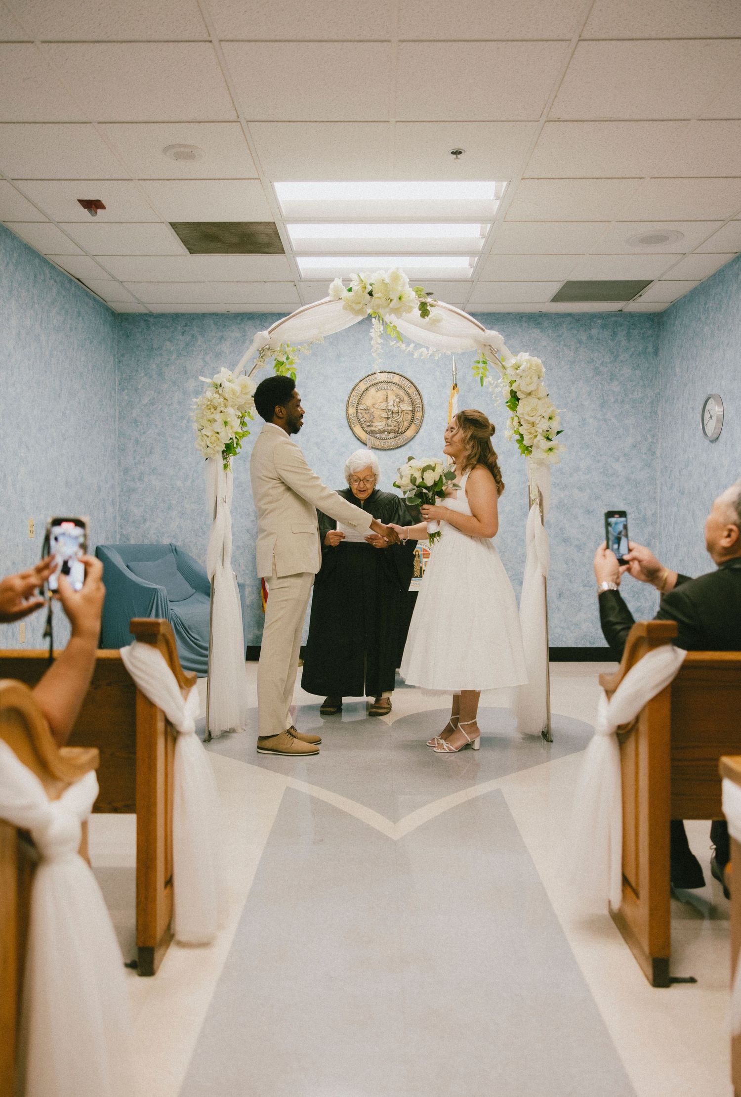Guests take photos during an intimate wedding ceremony under a floral arch.