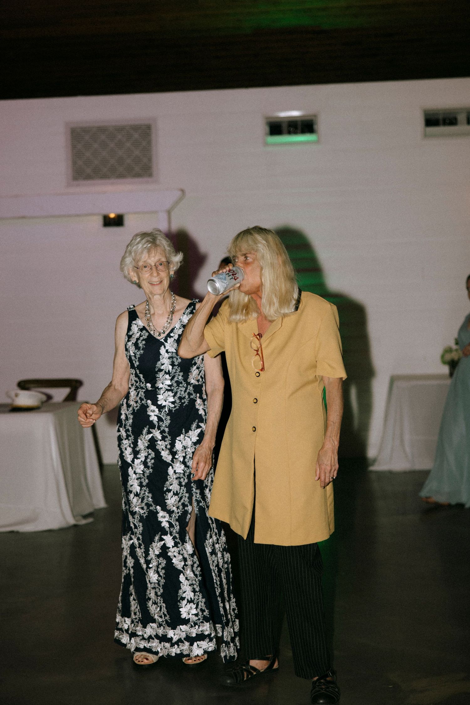 Two elderly women at a wedding reception, one in floral gown, other in yellow jacket drinking from a cup.