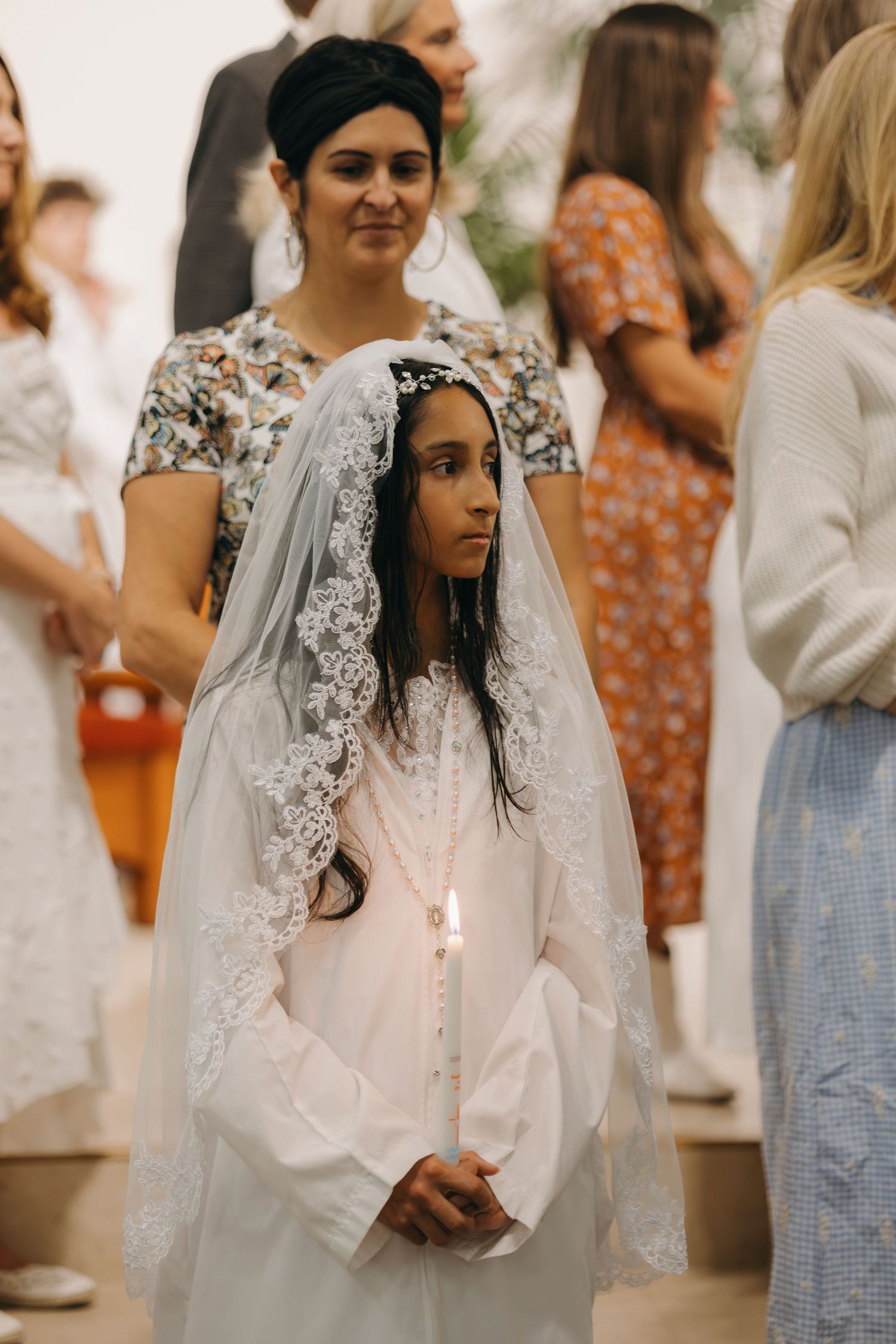 Young girl in white First Communion dress and lace veil holding lit candle during Catholic ceremony.
