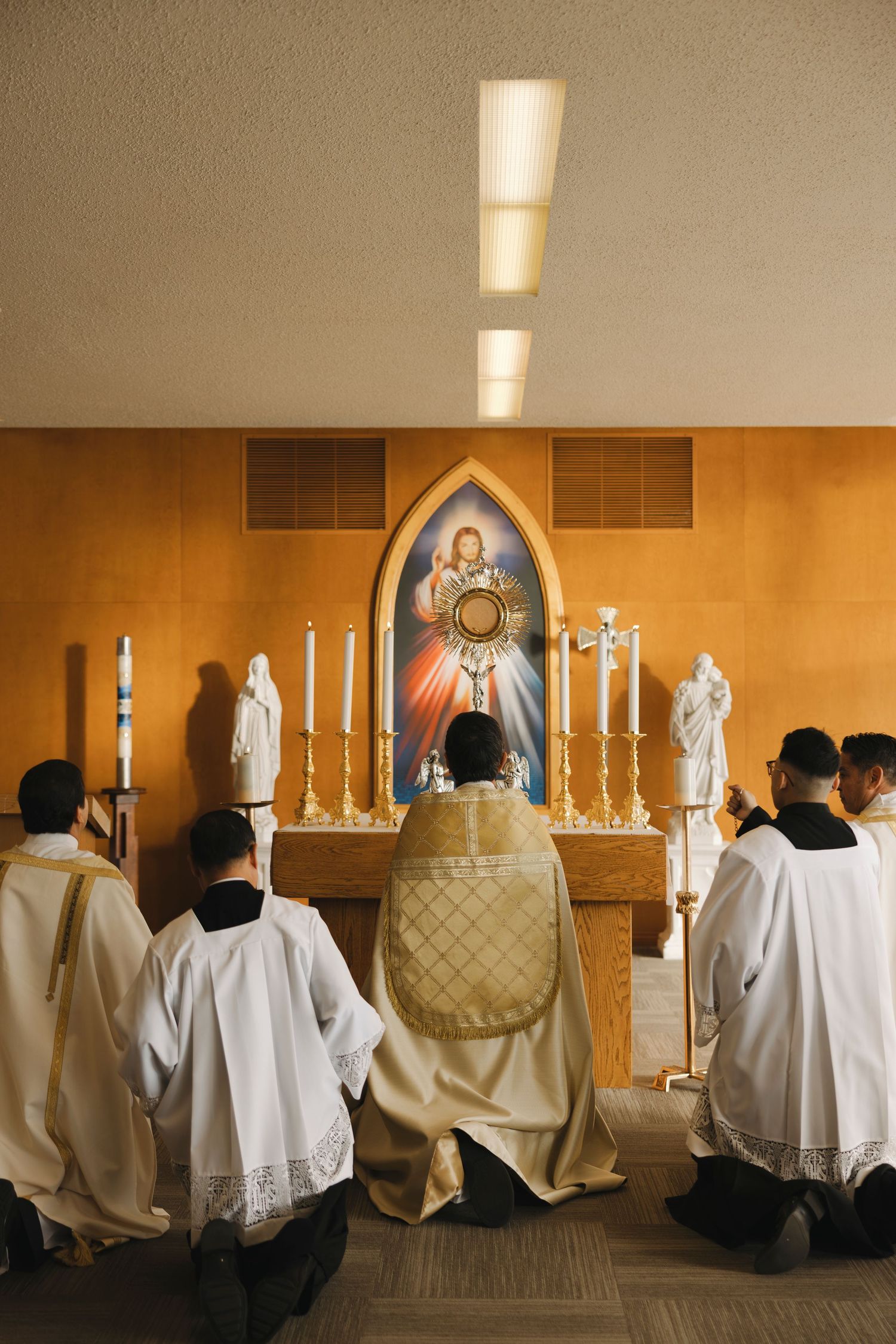 Clergy members in white robes kneeling before an altar with religious artwork and candles.