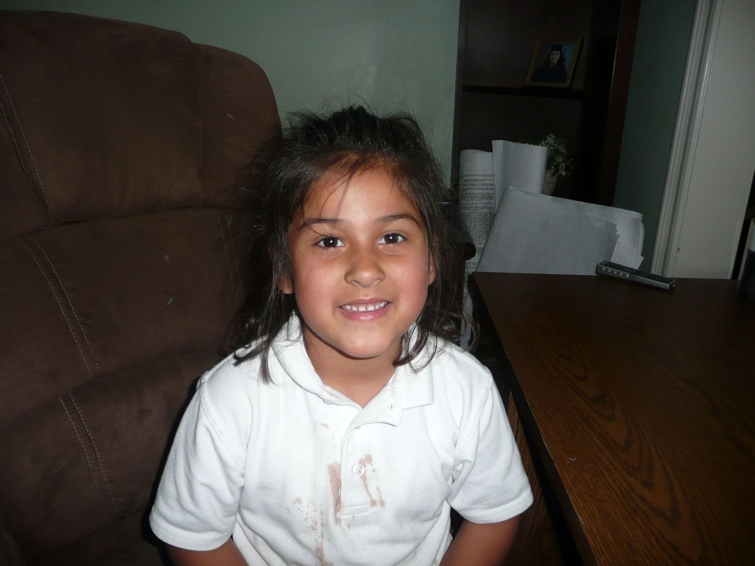 A young student in a white polo shirt smiles while sitting at a wooden table.