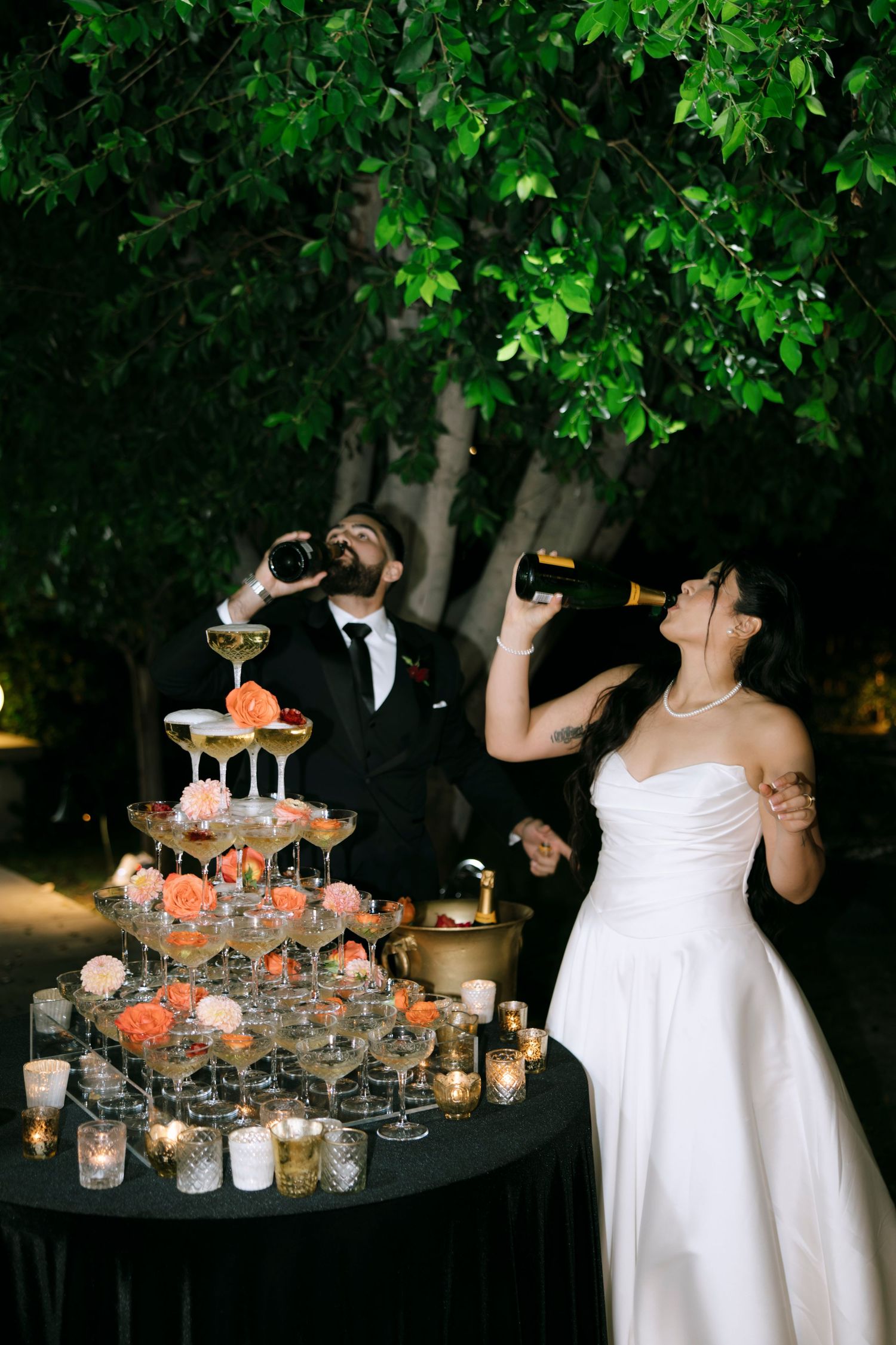 A couple toasts with champagne next to an illuminated tower of glasses at their wedding reception.