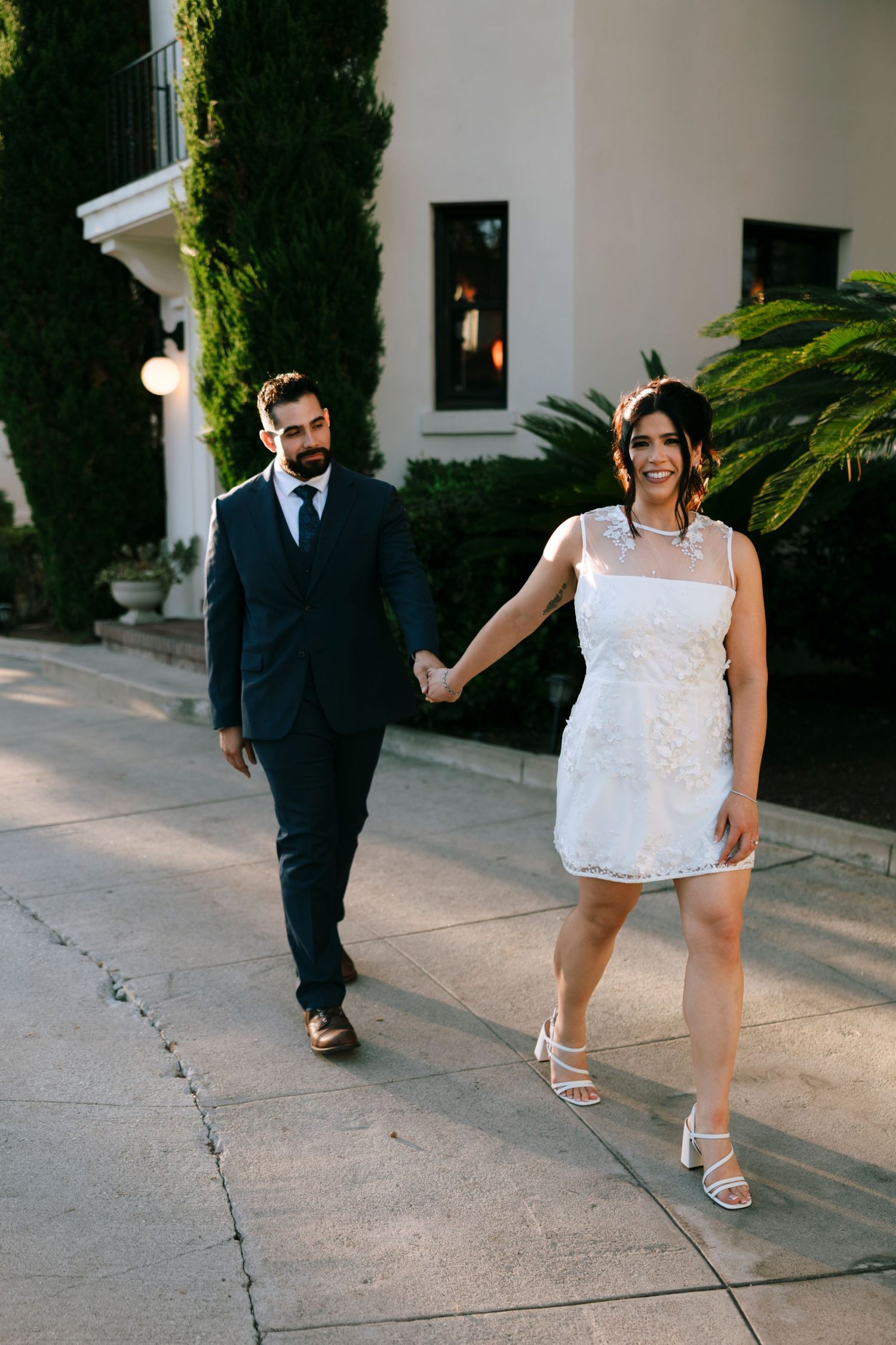 A couple playfully interacts on a concrete path outside a white building.