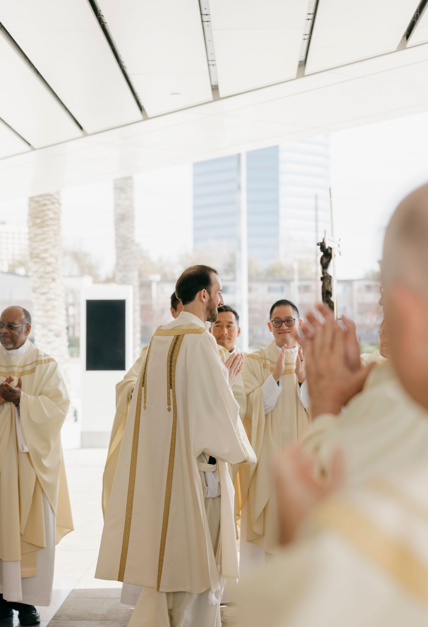 Religious ceremony with clergy in cream vestments standing against bright windows.
