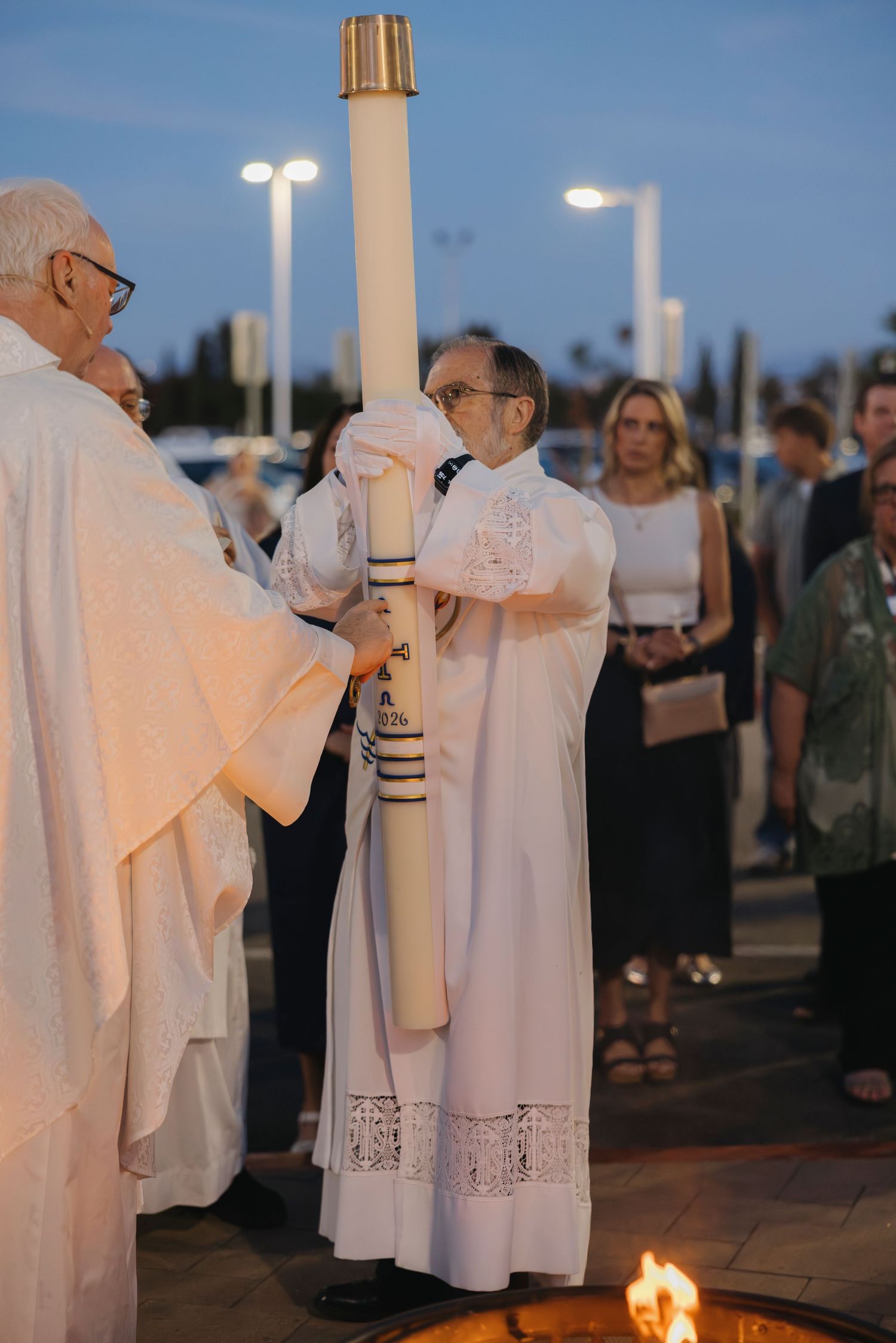 Deacon in white alb holds large Paschal candle during Catholic Easter Vigil liturgy outdoors.