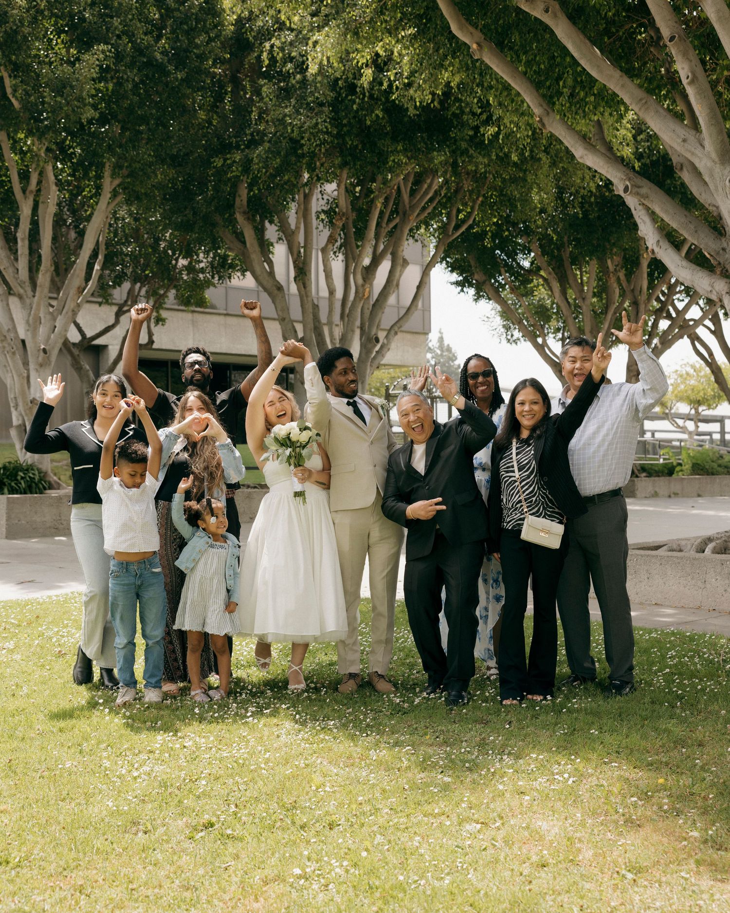 Wedding party and family members celebrate with raised arms outdoors under trees.