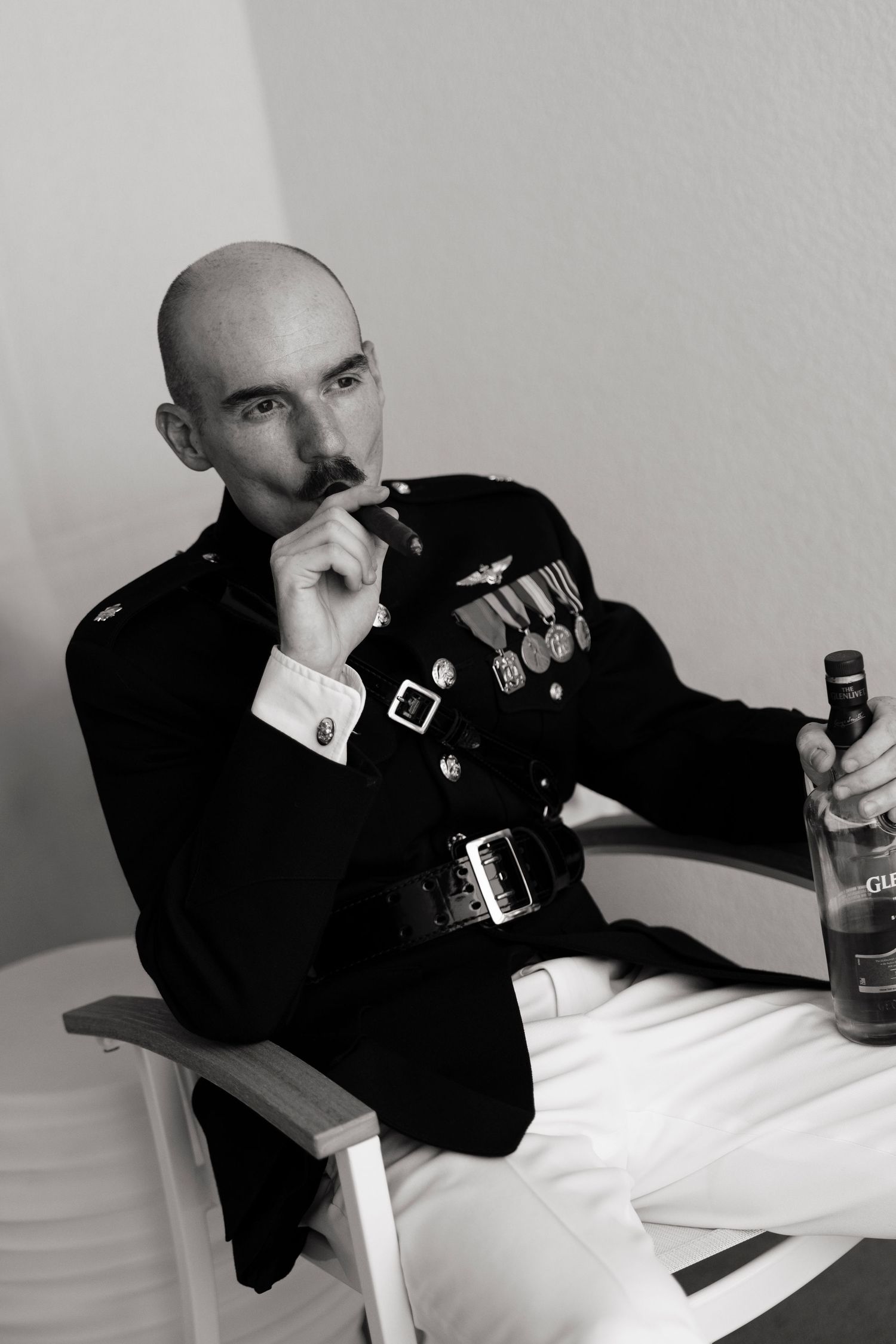 Bald man in military dress uniform with medals holds cigar and bottle, seated in director's chair, black and white photo.