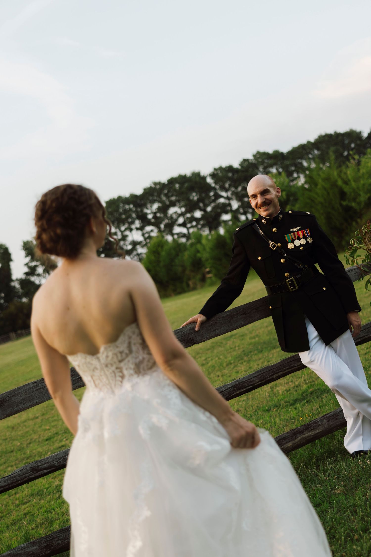 Marine groom in dress uniform smiles at bride in white wedding gown near rustic fence on green lawn.