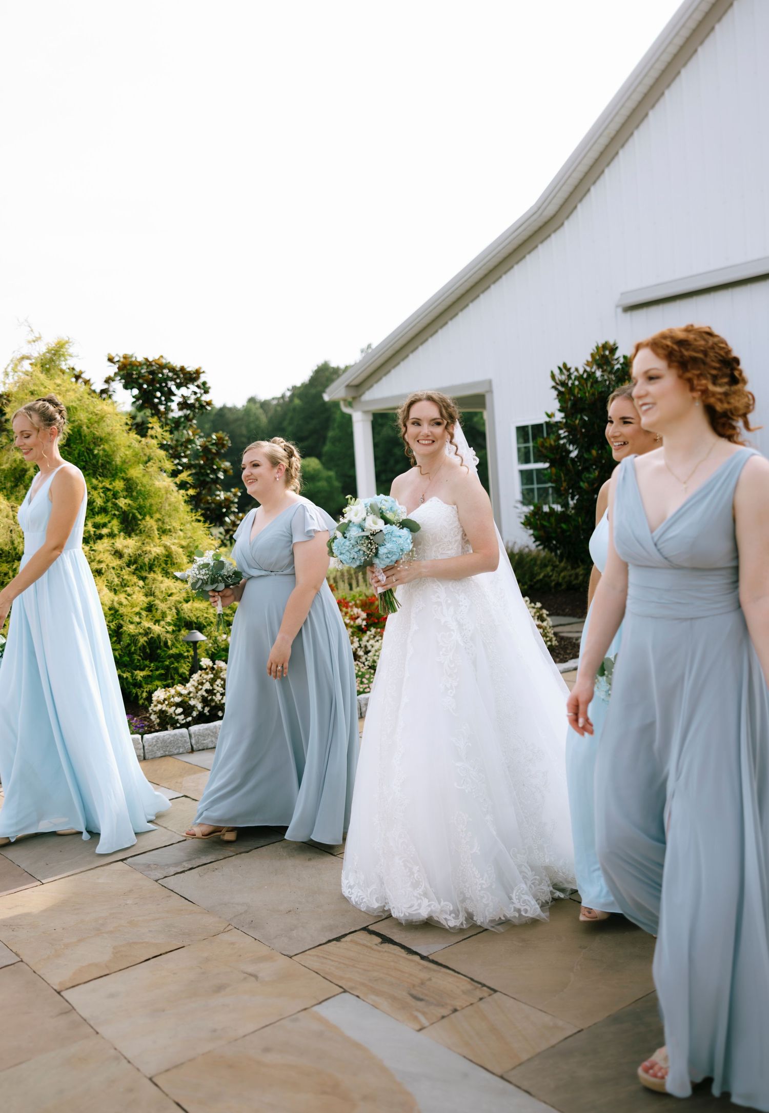 Bride in white lace gown walks with bridesmaids in light blue dresses outside a white venue.