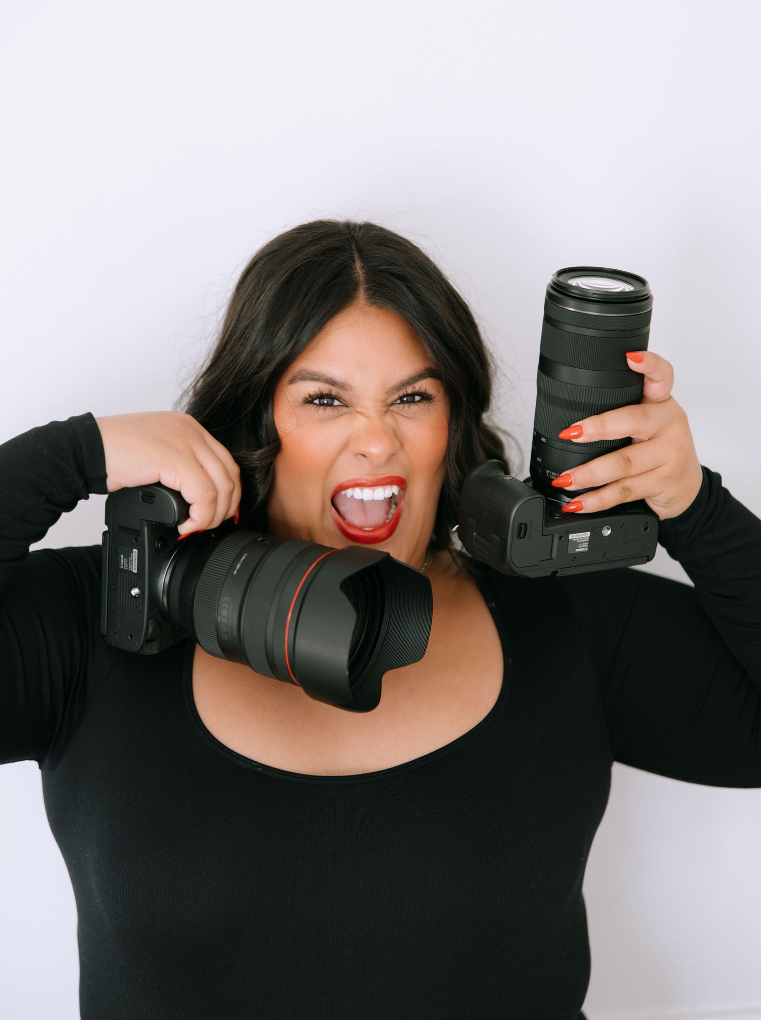 Woman in black top playfully holds two Canon DSLR cameras while making a funny face against white background.