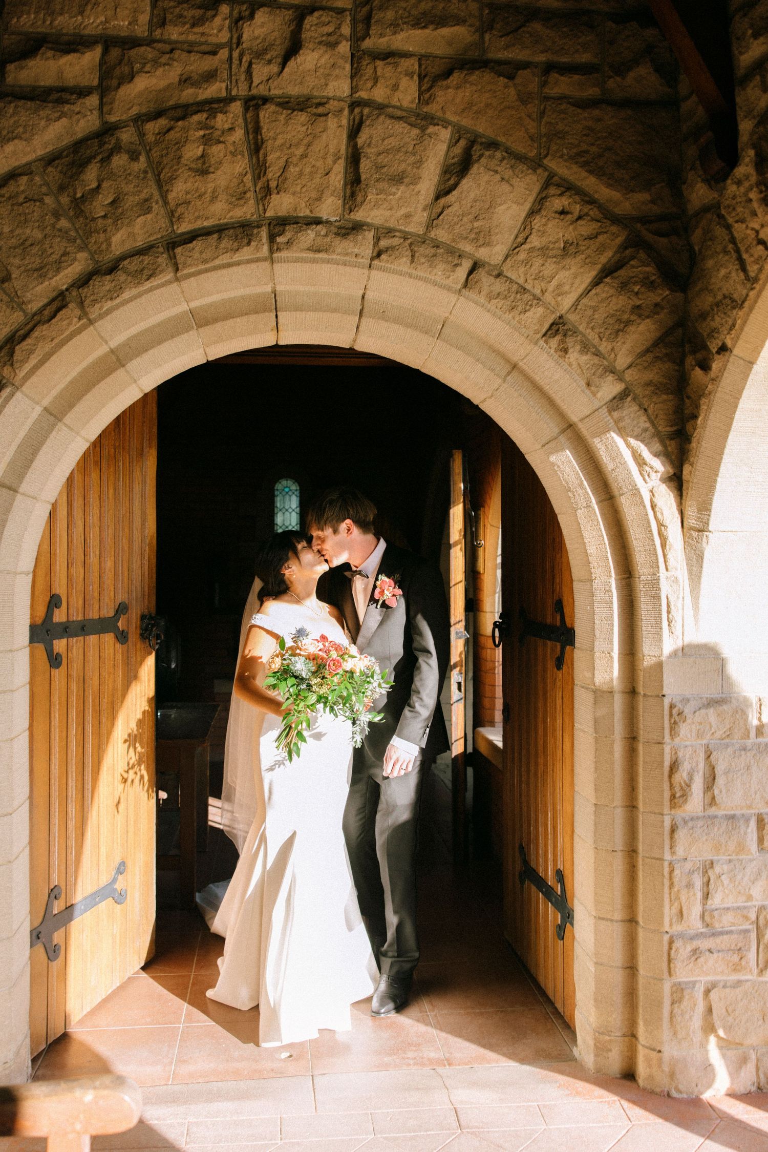 Bride and groom kissing under stone archway of church doors, bride holding floral bouquet in white wedding dress.