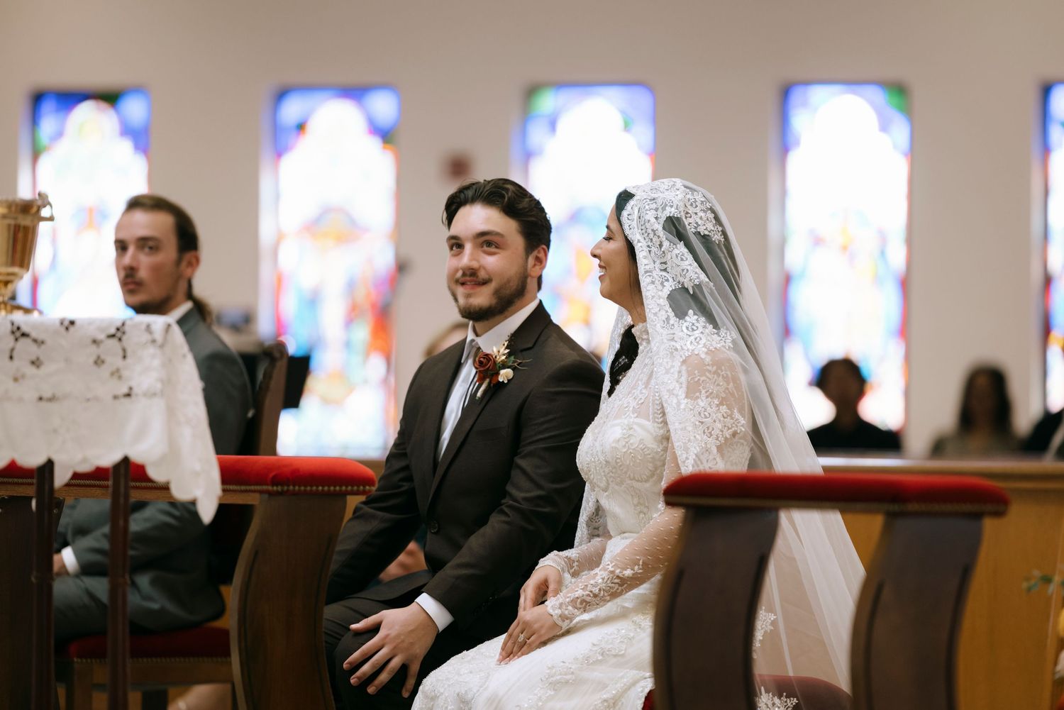 Stained glass windows illuminate a traditional church ceremony with clergy and attendees.