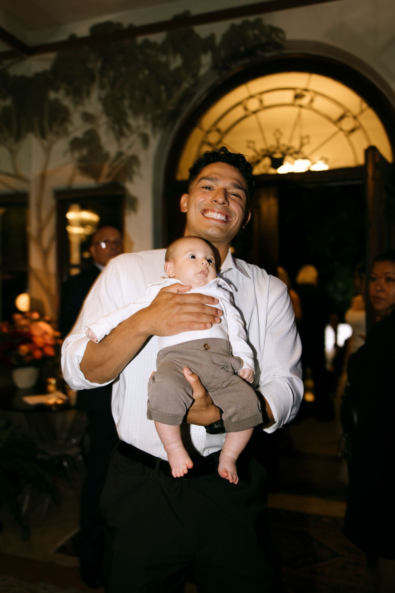 A tender moment at an evening celebration as a parent holds a baby dressed in grey at an elegant venue.