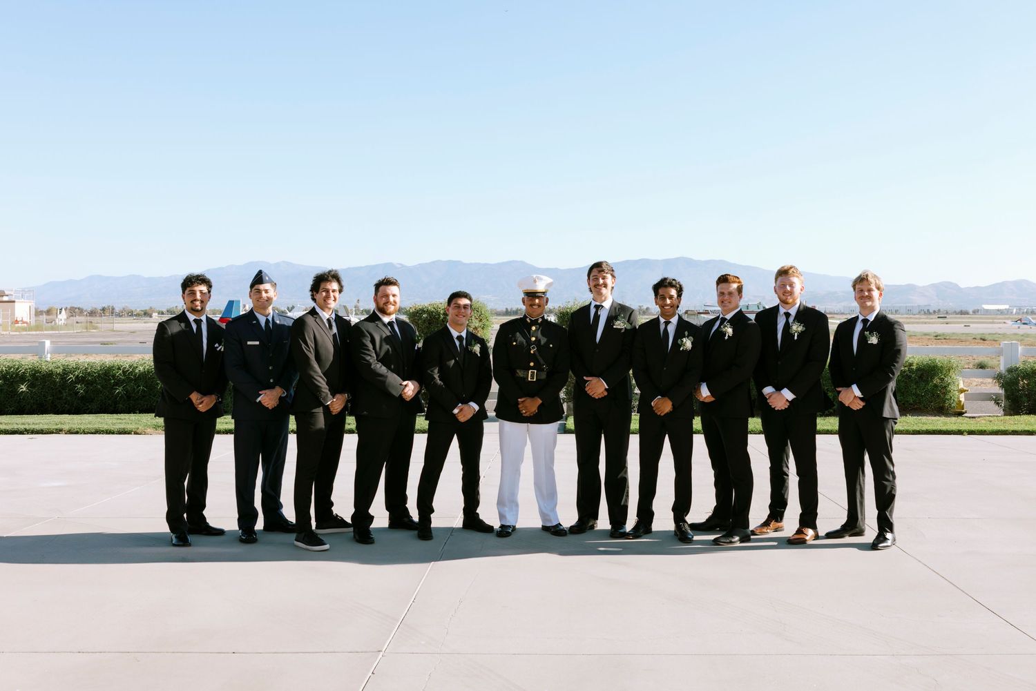 Groomsmen in black suits lined up with bride in white dress against mountain backdrop.