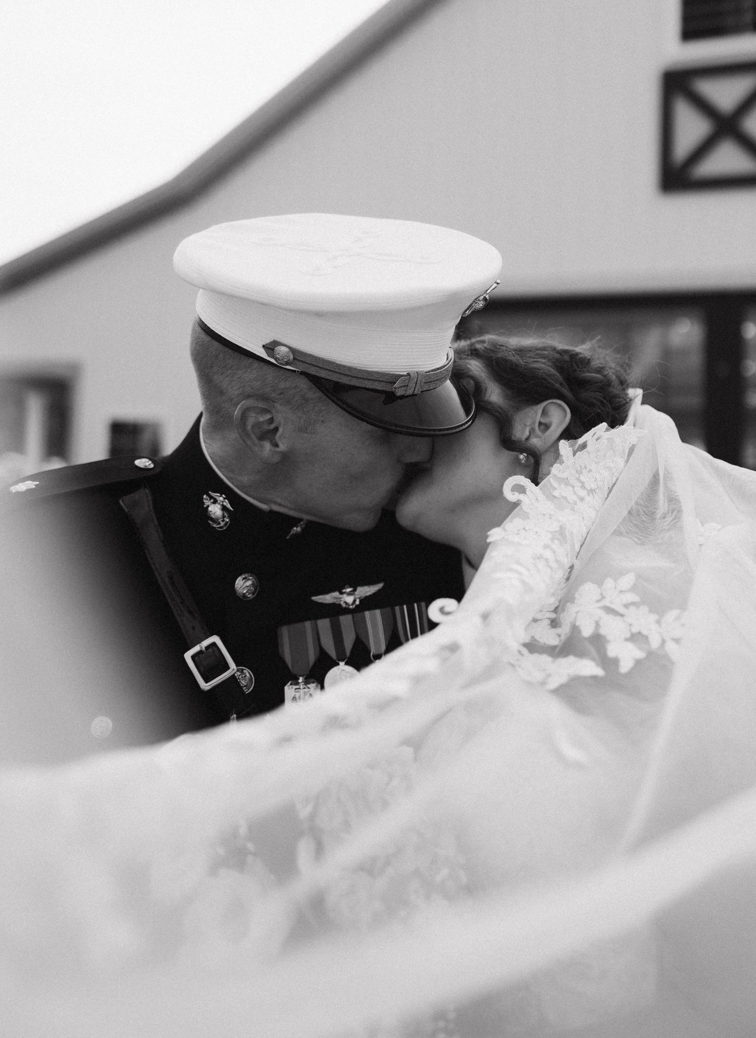 Black and white photo of a Marine in dress uniform sharing a kiss with his bride in a flowing lace veil.