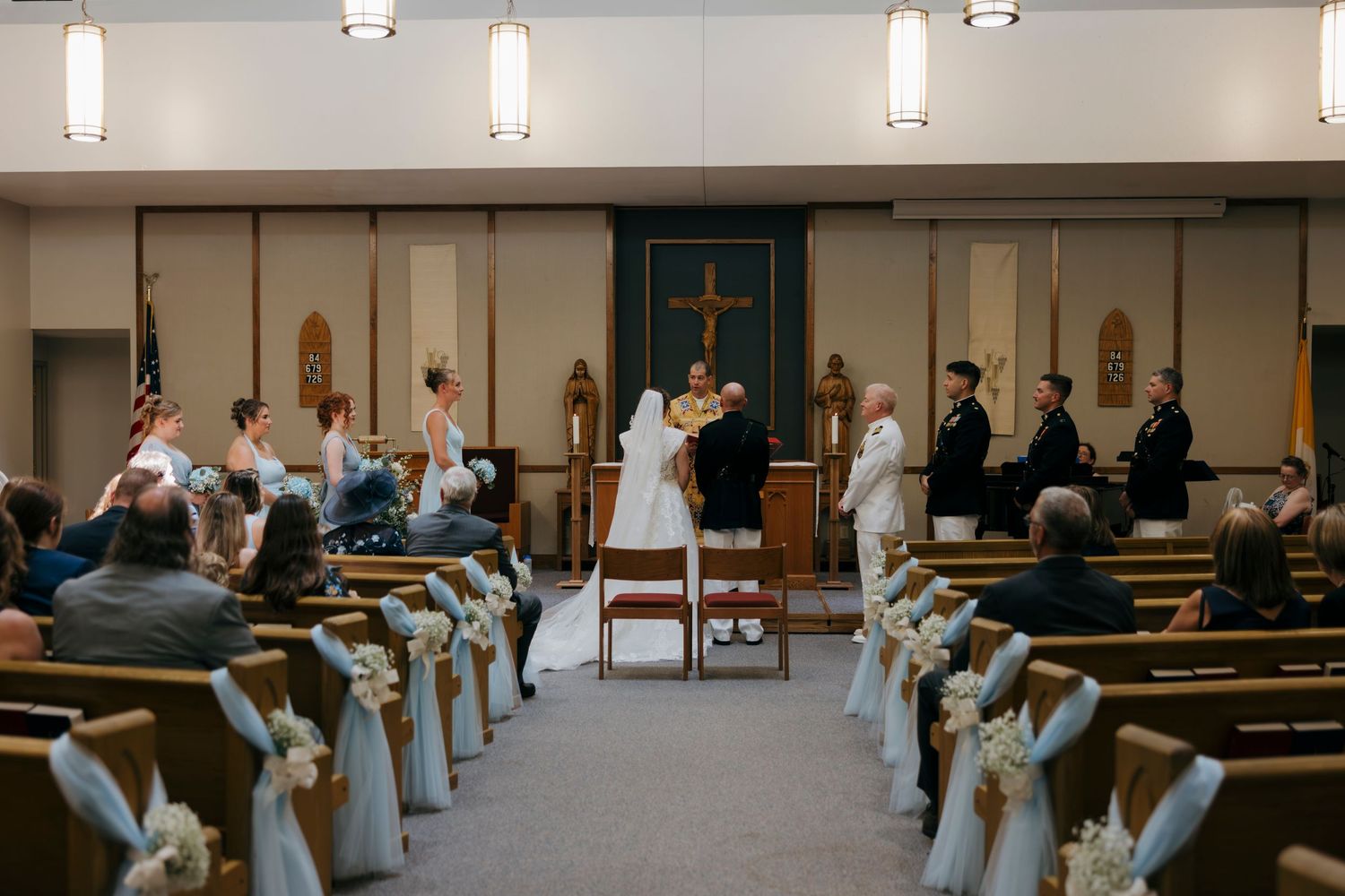 Elegant church wedding ceremony with bride and groom at altar, decorated pews with white florals, guests seated in wooden benches.
